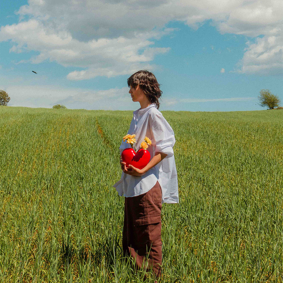 Woman in a green field wearing brown pants and white shirt holding red ceramic DOIY love vase filled with yellow flowers
