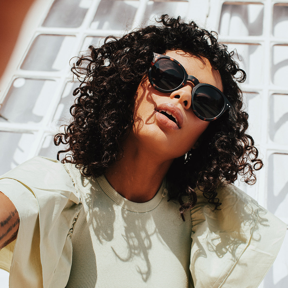 Woman with brown curly hair wearing Izipizi shape m tortoise sunglasses in front of lattice wall