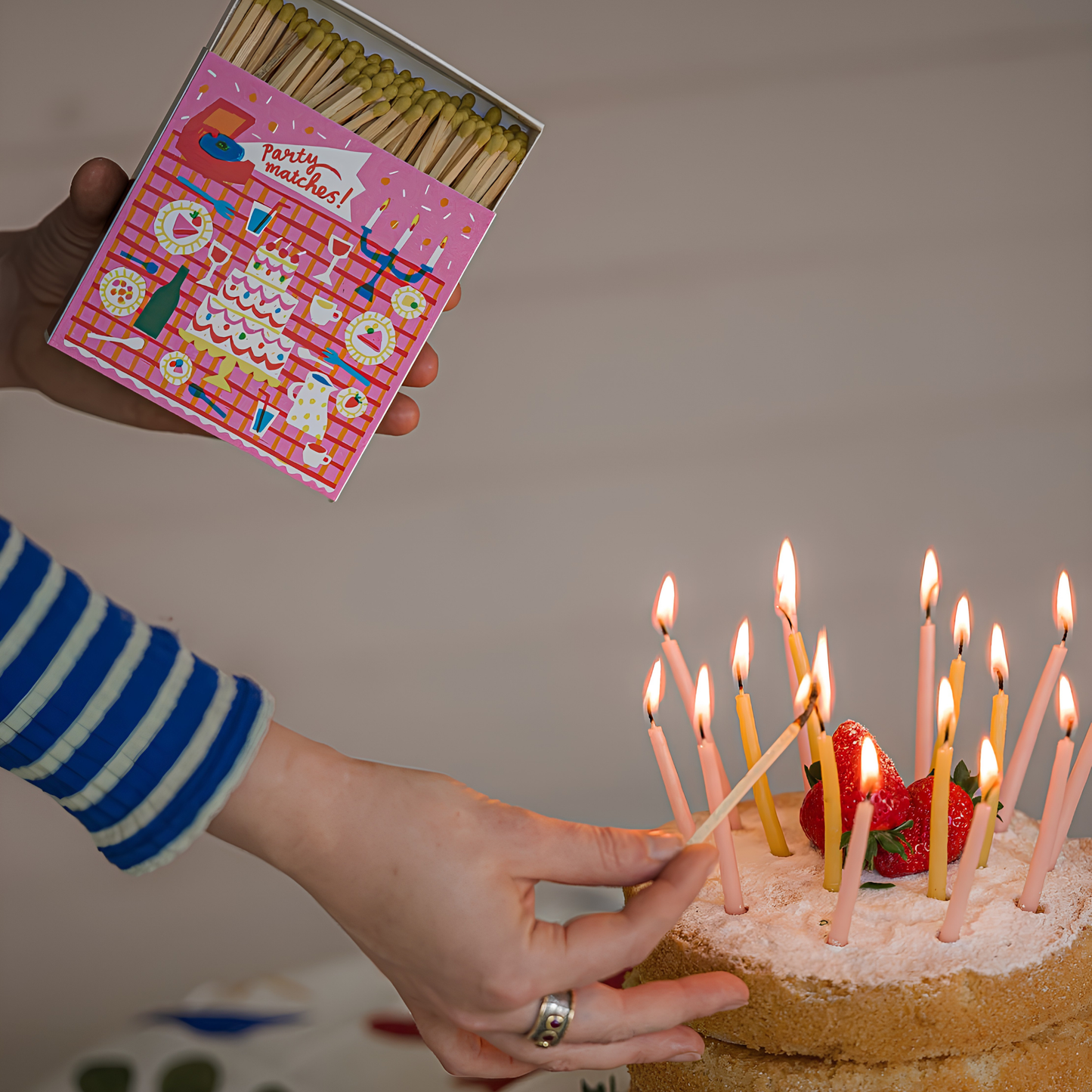 Close up on a woman lighting the candles on a cake using the Archivist Luxury Matches featuring an illustration Picnic Party with cakes and drinks, 'Party Matches' text.