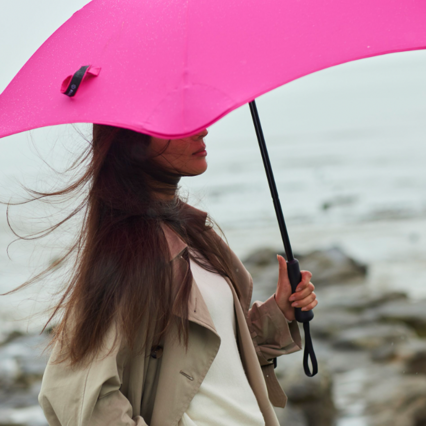 Woman holding open pink classic Blunt umbrella by rocks and water
