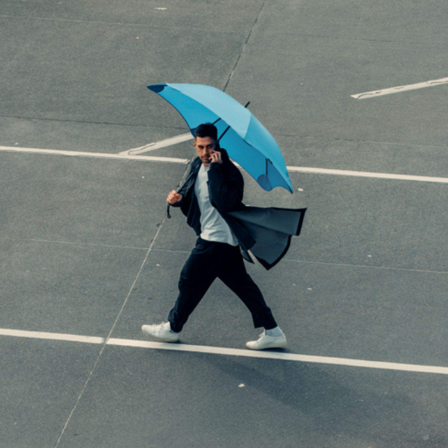 Man crossing street on phone holding open Blunt sky blue classic umbrella over shoulder