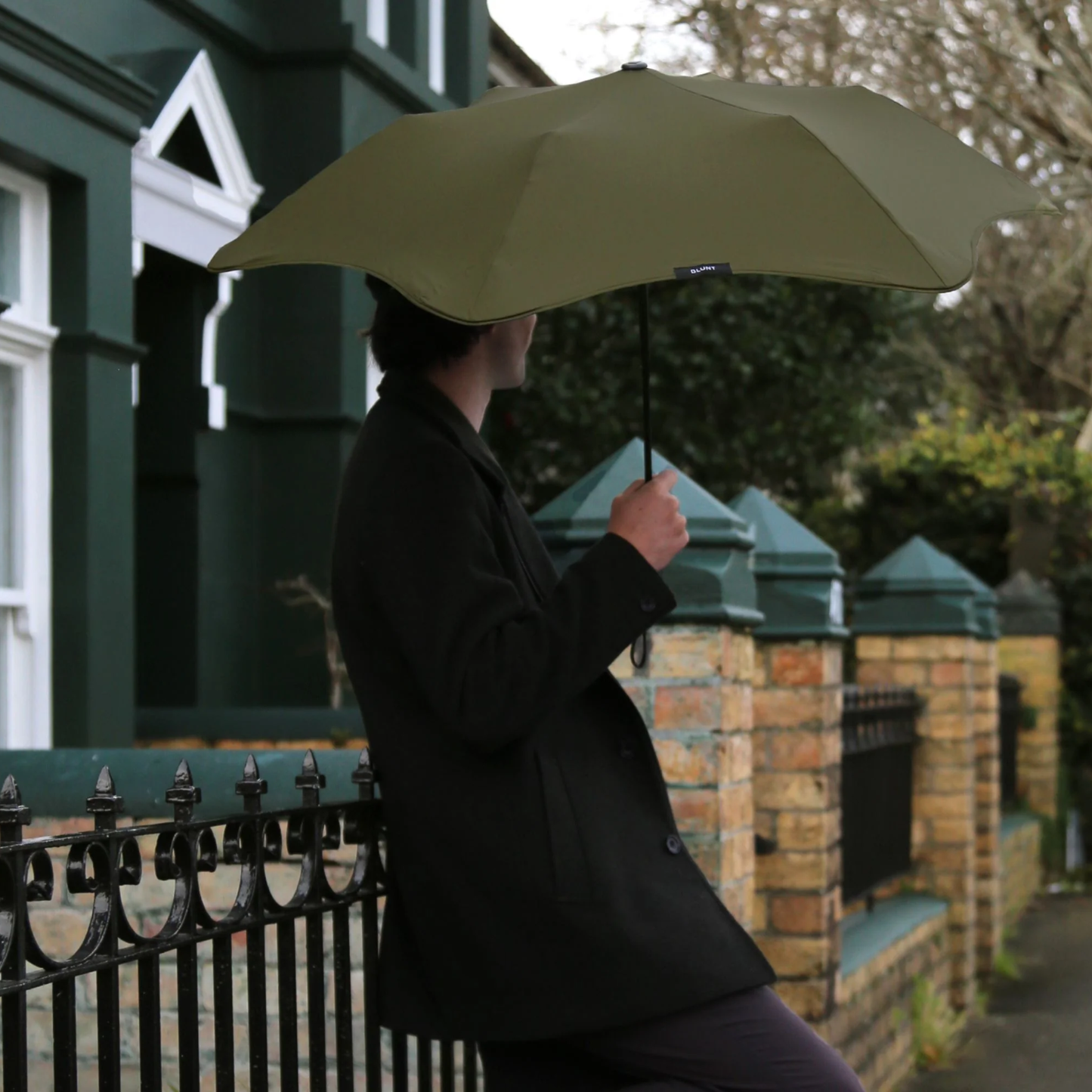 A man standing and leaning to a fence while holding the Blunt Umbrella Metro Moss Green, a timeless, compact, collapsible and convenient, consisting of the detachable UV fabric canopy, sturdy frame, shaft and handle. Fits in your handbag, tote or backpack. Built for fast living, those who travel light, and any weather.