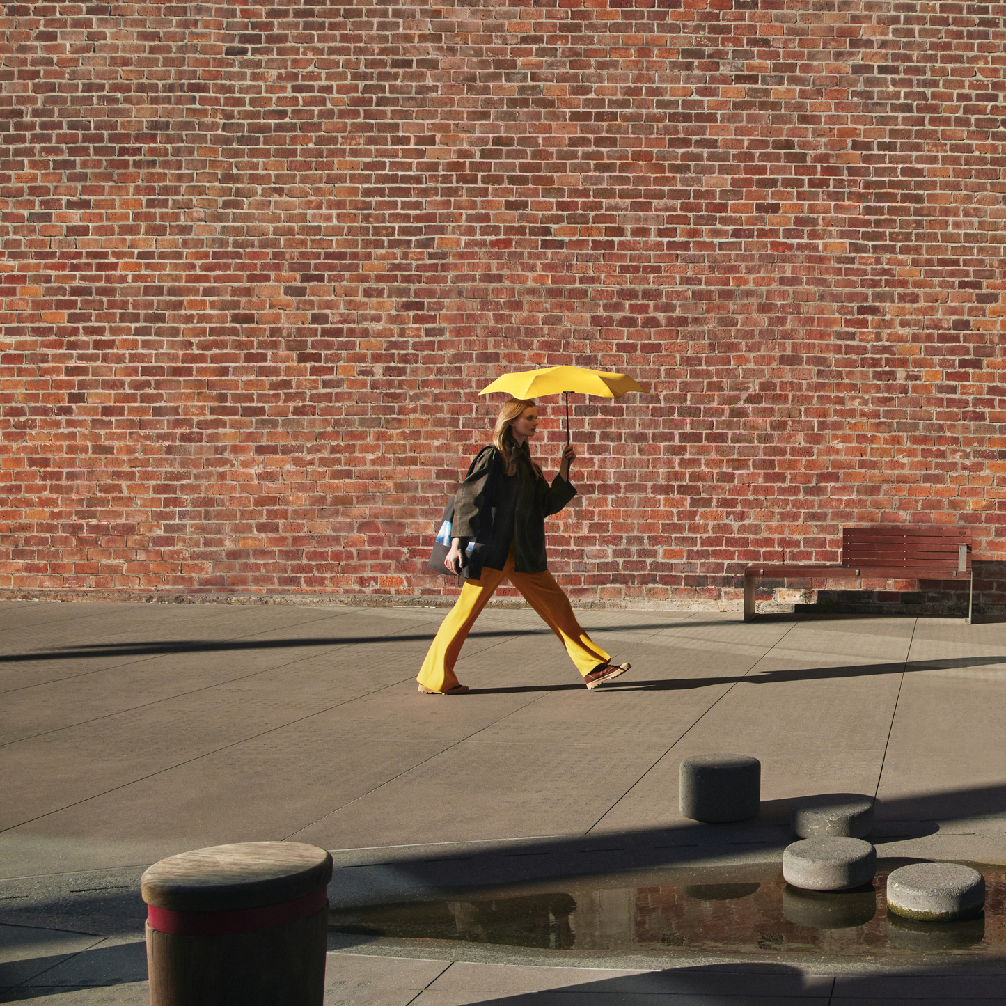 A woman walking holding the open Blunt Umbrella Neo Citrus Yellow at 98cm, featuring a compact design with high tensioned canopy, wind tested construction and recycled rPET fabric for rain or shine.