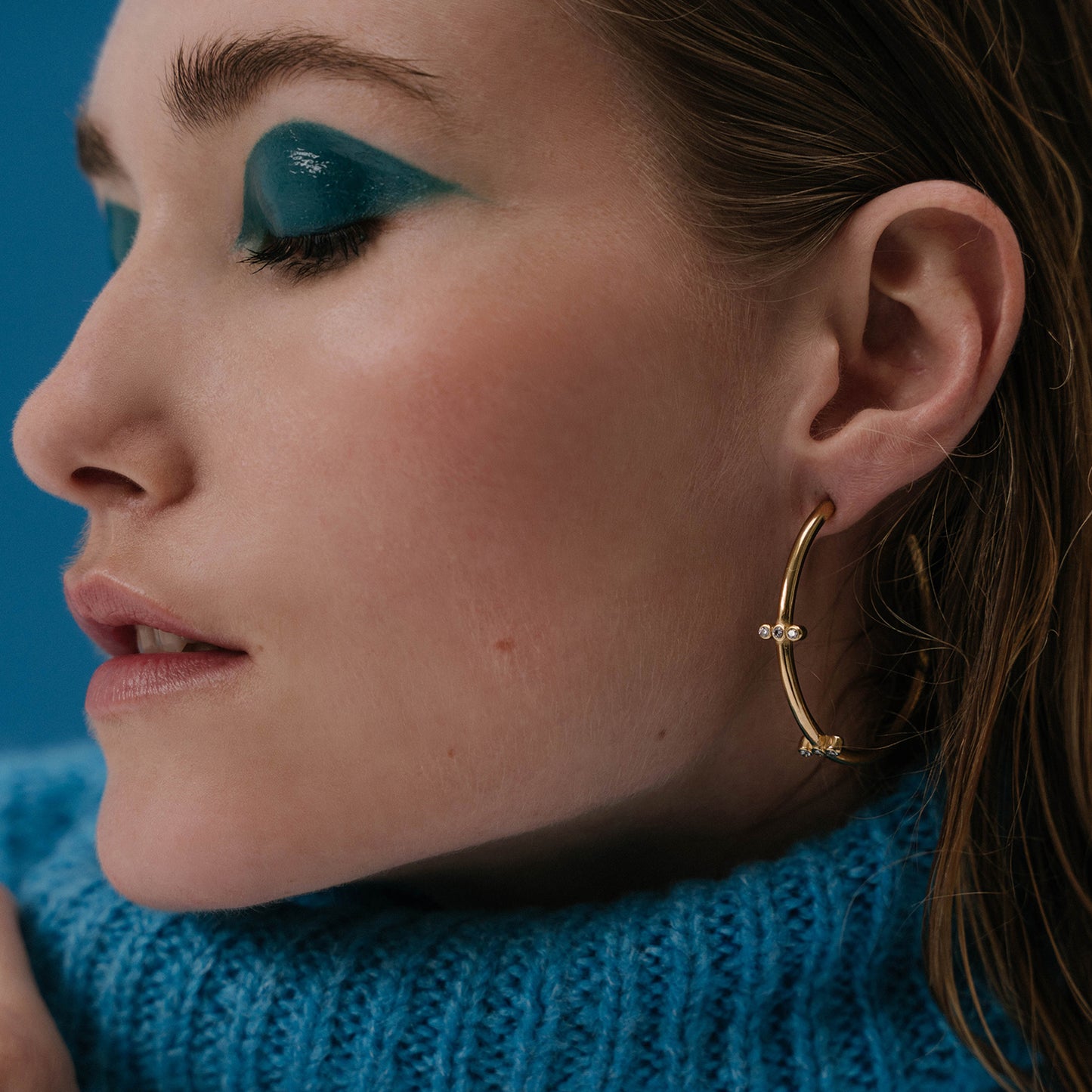 A woman wearing on her ear the AEC Paris Aura White Zirconium Oxide earrings - multi-stone open hoop earrings with white cubic zirconia on 14k gold-plated stainless steel, hypoallergenic, durable, elegant everyday jewellery, 45mm diameter.