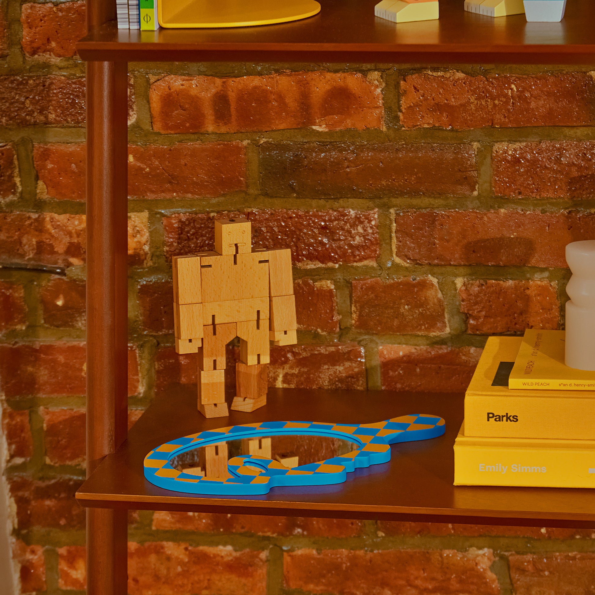 Areaware Cubebot Micro wooden toy robot on a wooden rack with books and handheld mirror - natural beech-wood figure with elastic joints, folds into a 1.5-inch cube and poses in multiple positions; compact, durable, and suitable for ages 4+.