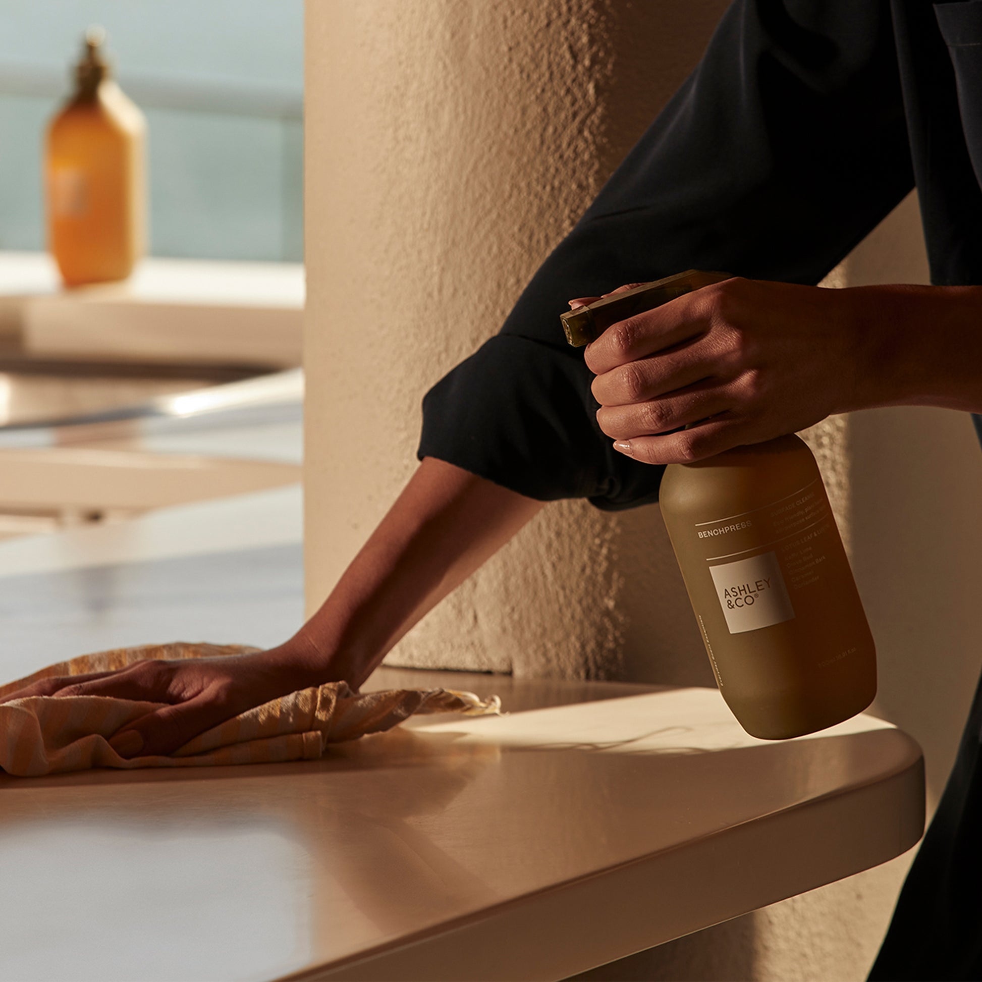 A woman cleaning a surface using Ashley and Co 'Benchpress' surface spray in lotus leaf & lustre fragrance.
