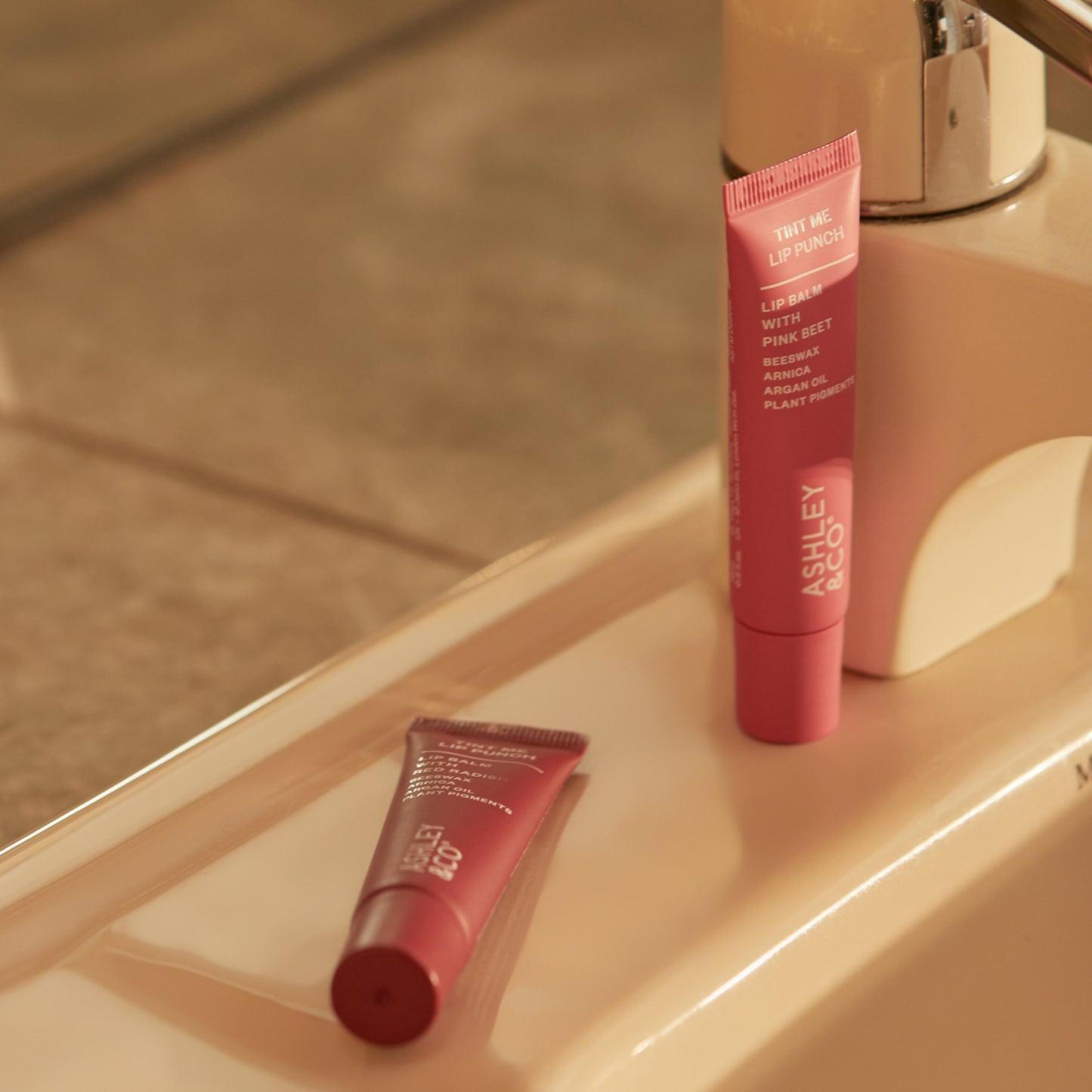 Ashley and Co 'Tint Me Lip Punch' pink coloured lip balm. on a bathroom counter.