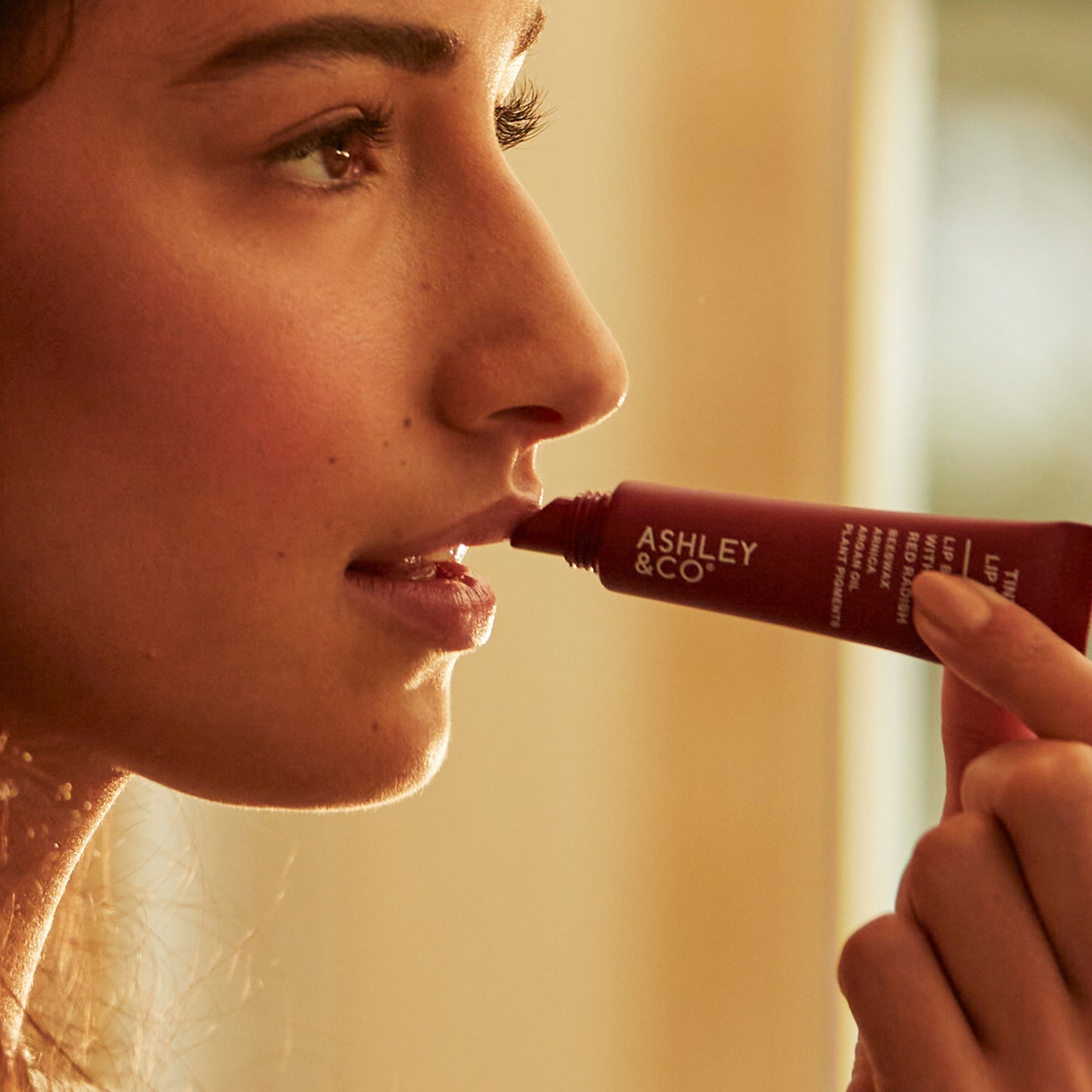 A woman applying Ashley and Co 'Tint Me Lip Punch' beet red coloured lip balm.