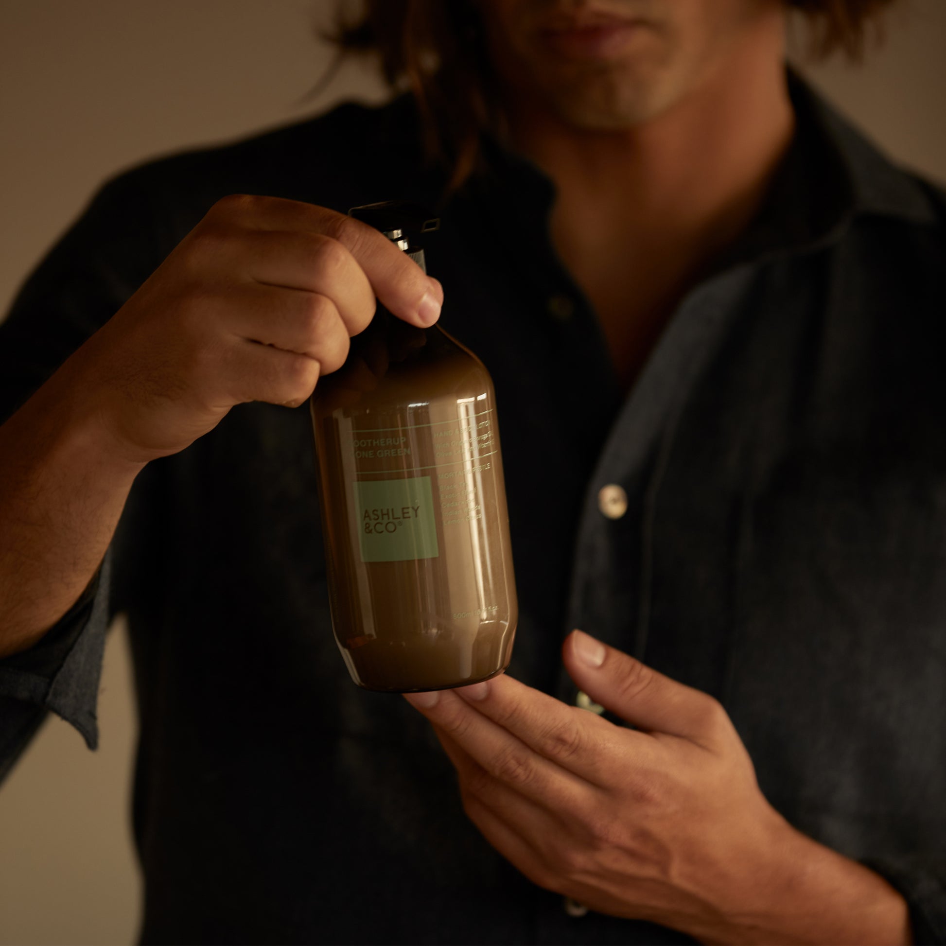 A man holding a bottle of Ashley and Co 'Sootherup Gone Green' hand and body lotion in mortar and pestle fragrance.