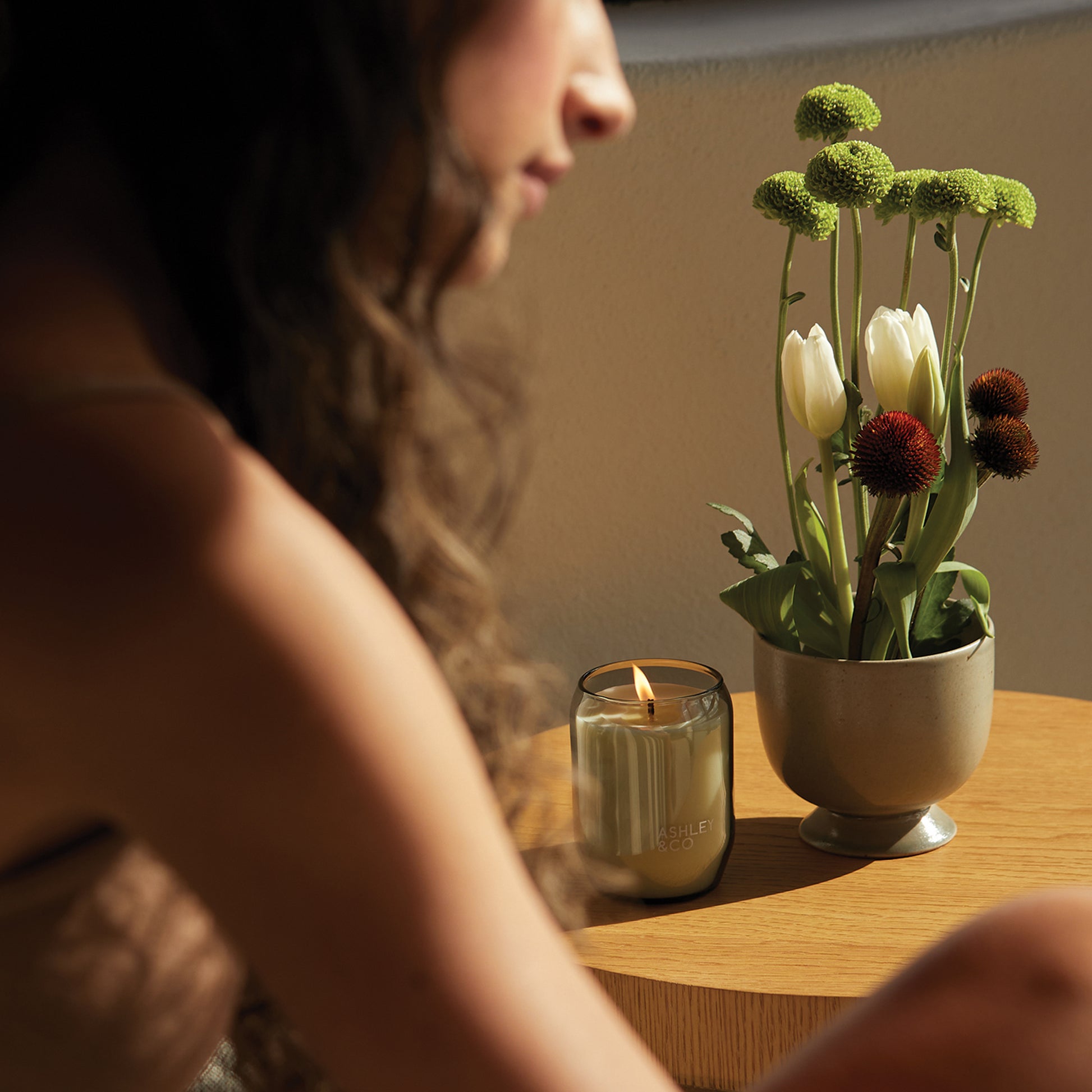 An Ashley and Co 'Waxed Perfume' scented candle in blossom and gilt fragrance on a side table next to a vase of flowers and with a woman sitting next to it.
