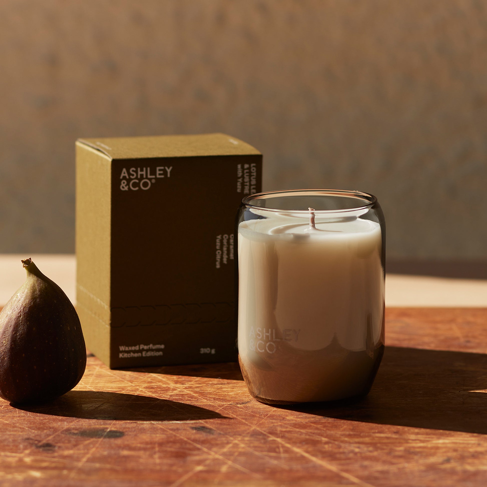 Ashley and Co 'Waxed Perfume' scented candle in lotus leaf and lustre fragrance on a counter next to a fig.