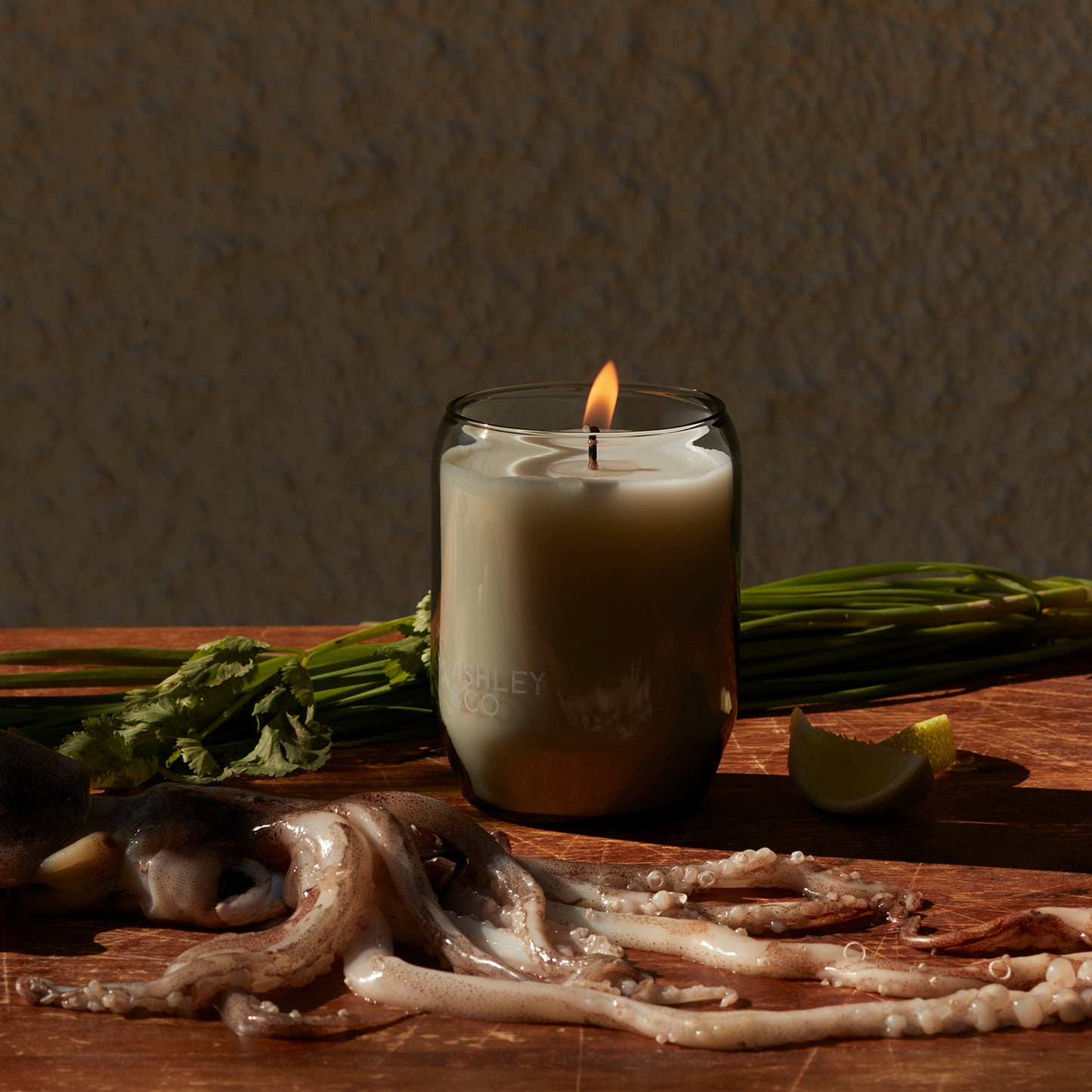 Ashley and Co 'Waxed Perfume' scented candle in lotus leaf and lustre fragrance on a kitchen counter next to squid and vegetables.