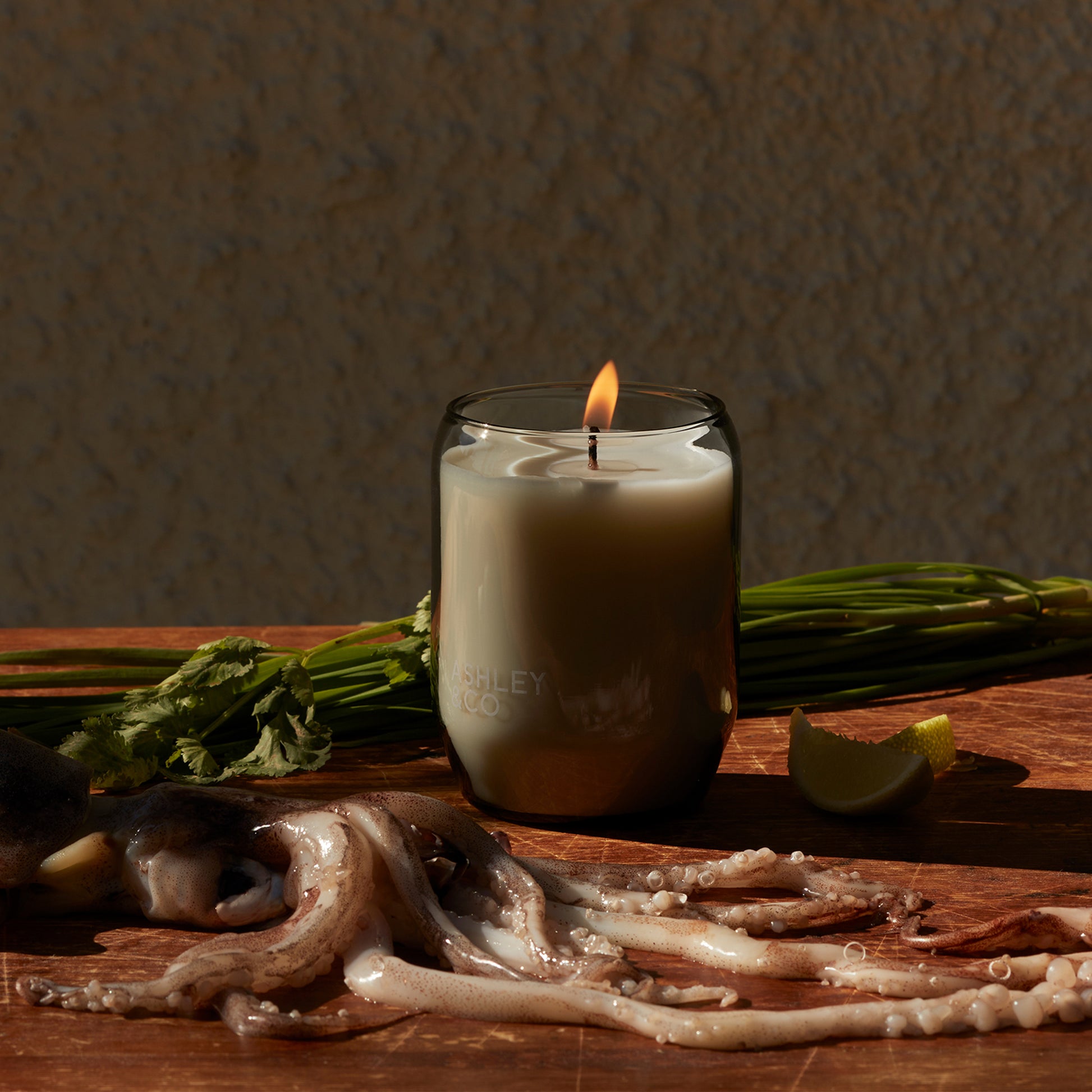 Ashley and Co 'Waxed Perfume' scented candle in lotus leaf and lustre fragrance on a kitchen counter next to squid and vegetables.