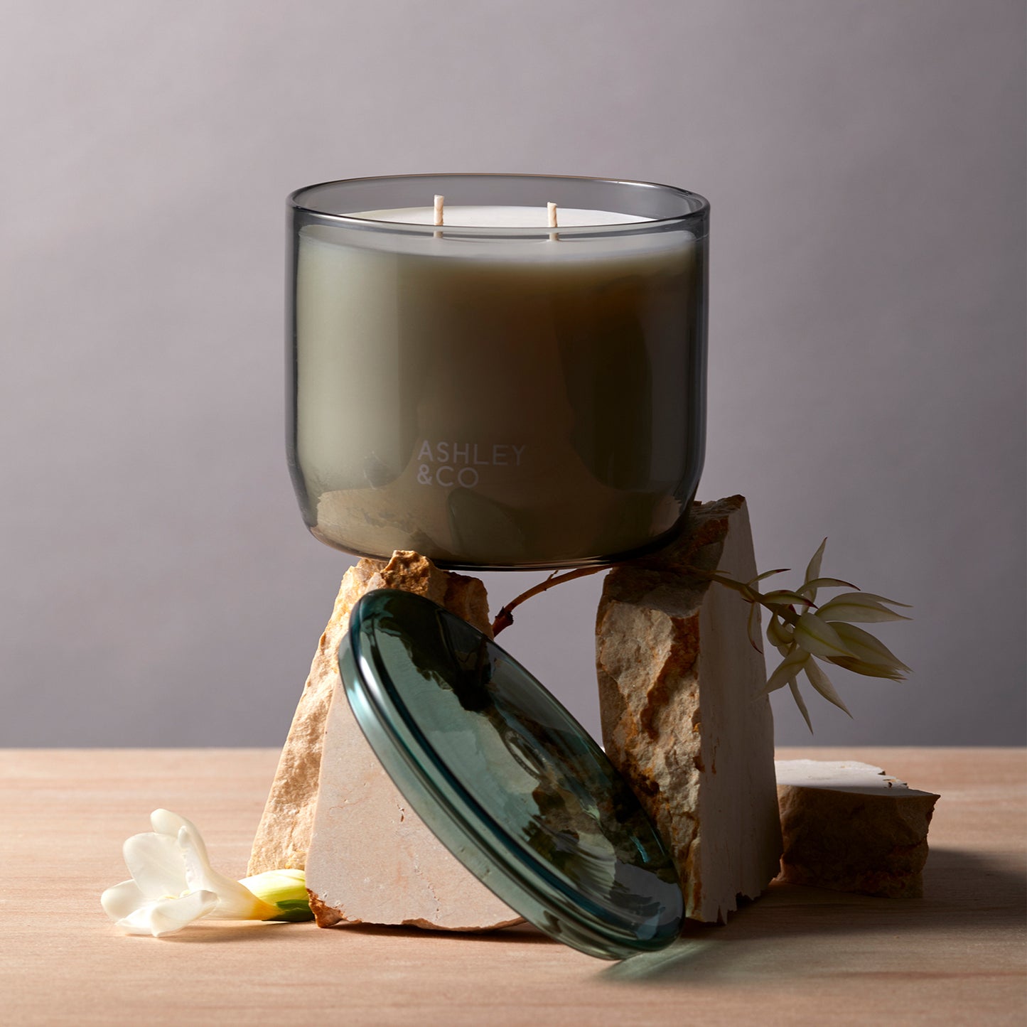 Ashley and Co 'Waxed Perfume' scented candle in blossom and gilt fragrance resting on shaeds of travertine stone, showing off the grey vessel and green lid.