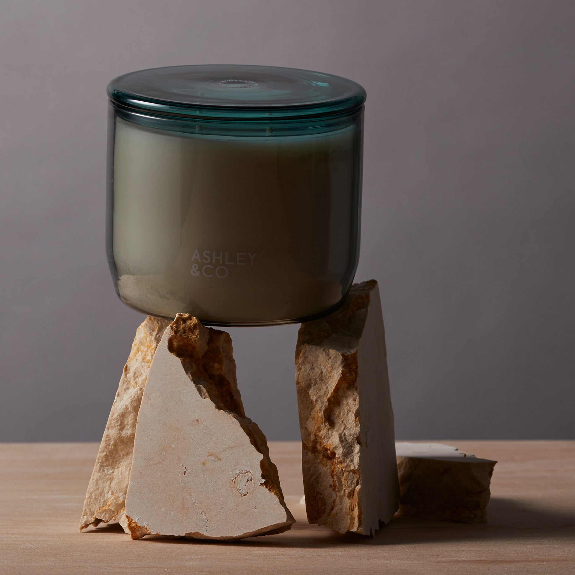 Ashley and Co 'Waxed Perfume' scented candle in tui and kaili fragrance resting on shaeds of travertine stone, showing off the grey vessel and green lid.