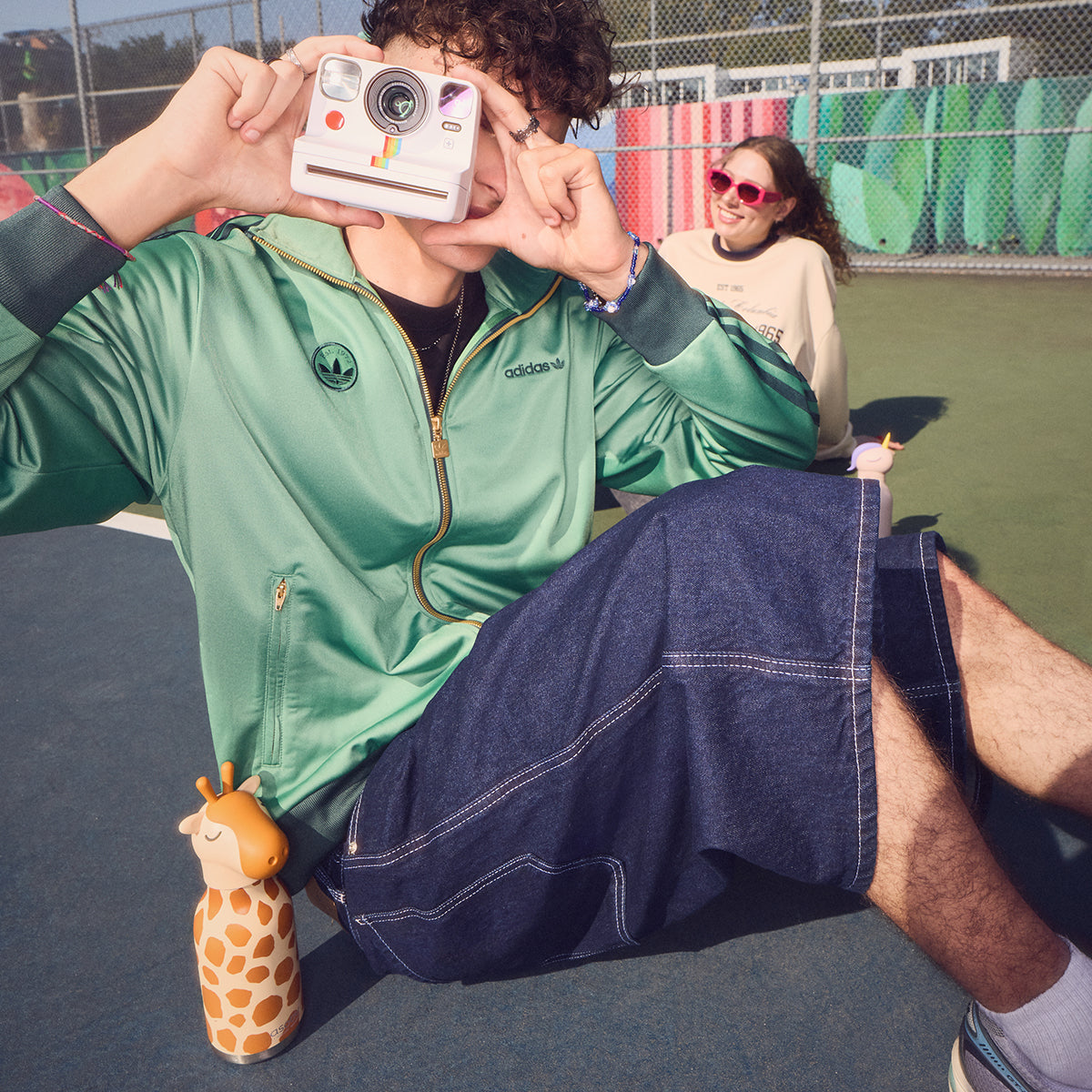 Bestie Bottle Giraffe on a tennis court next to a boy takng a polaroid photo,