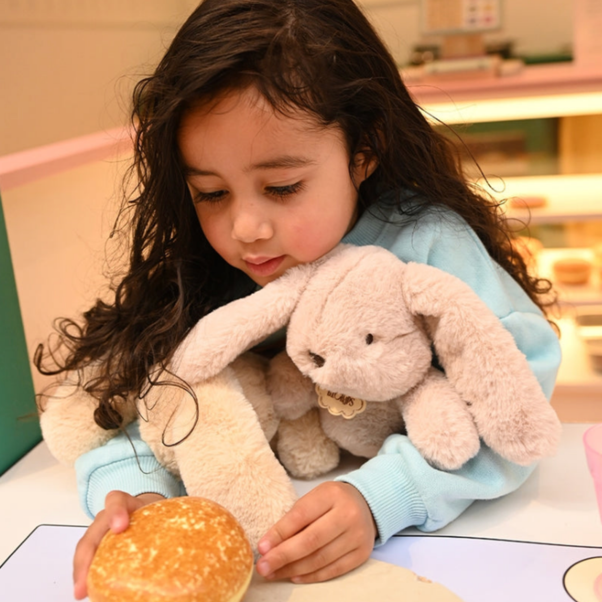 Small girl holding Bon Ton Toys Bubbles Bunny ballet pink plush toy in her hand eating a doughnut lifestyle shot
