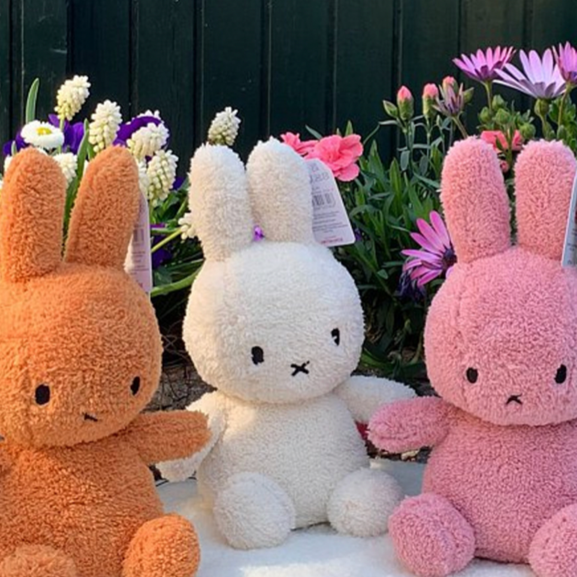 Three orange, cream and pink terry Bon Ton Toys Sitting Miffys sitting with flowers outside lifestyle shot