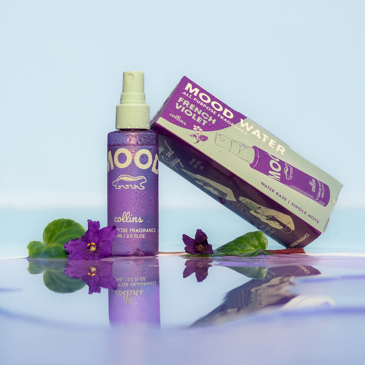 A travel sized bottle of french violet scented mood water by collins, placed next to its box on a reflective surface with violets.