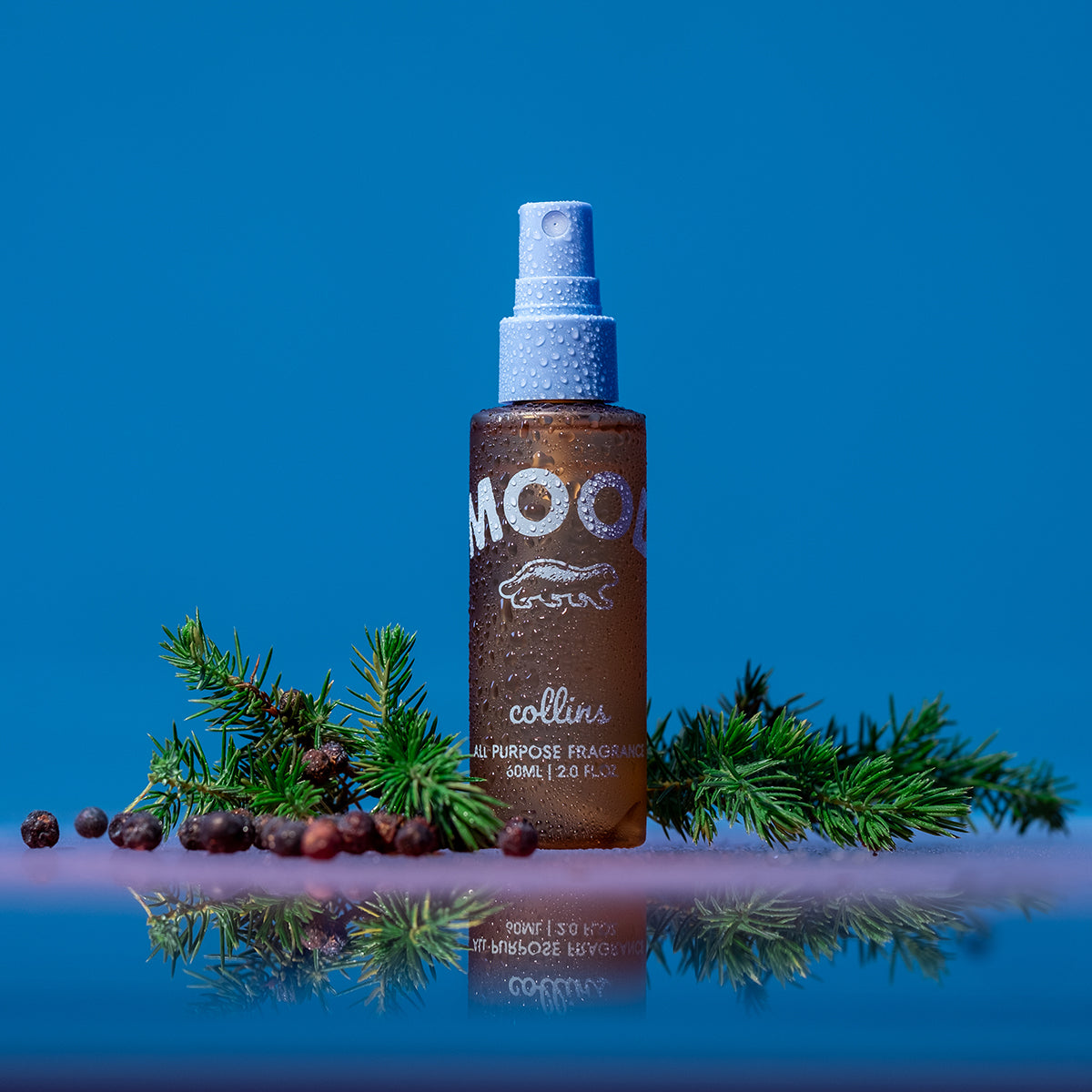 A travel sized bottle of scottish juniper scented mood water by collins placed on a reflective surface with juniper leaves and berries.