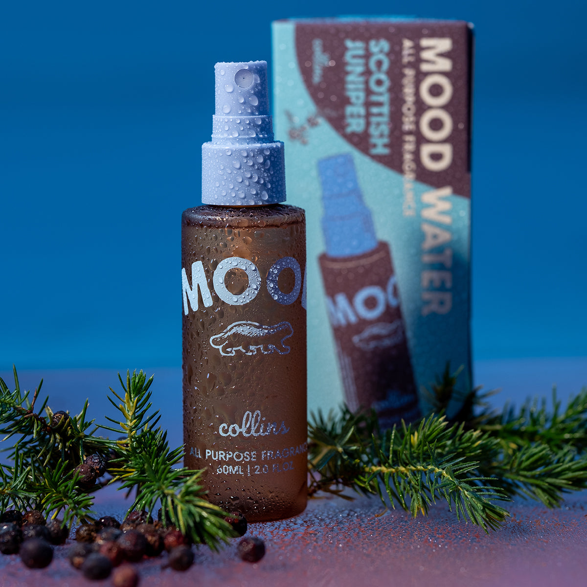 A travel sized bottle of scottish juniper scented mood water by collins placed with its box on a reflective surface with juniper leaves and berries.