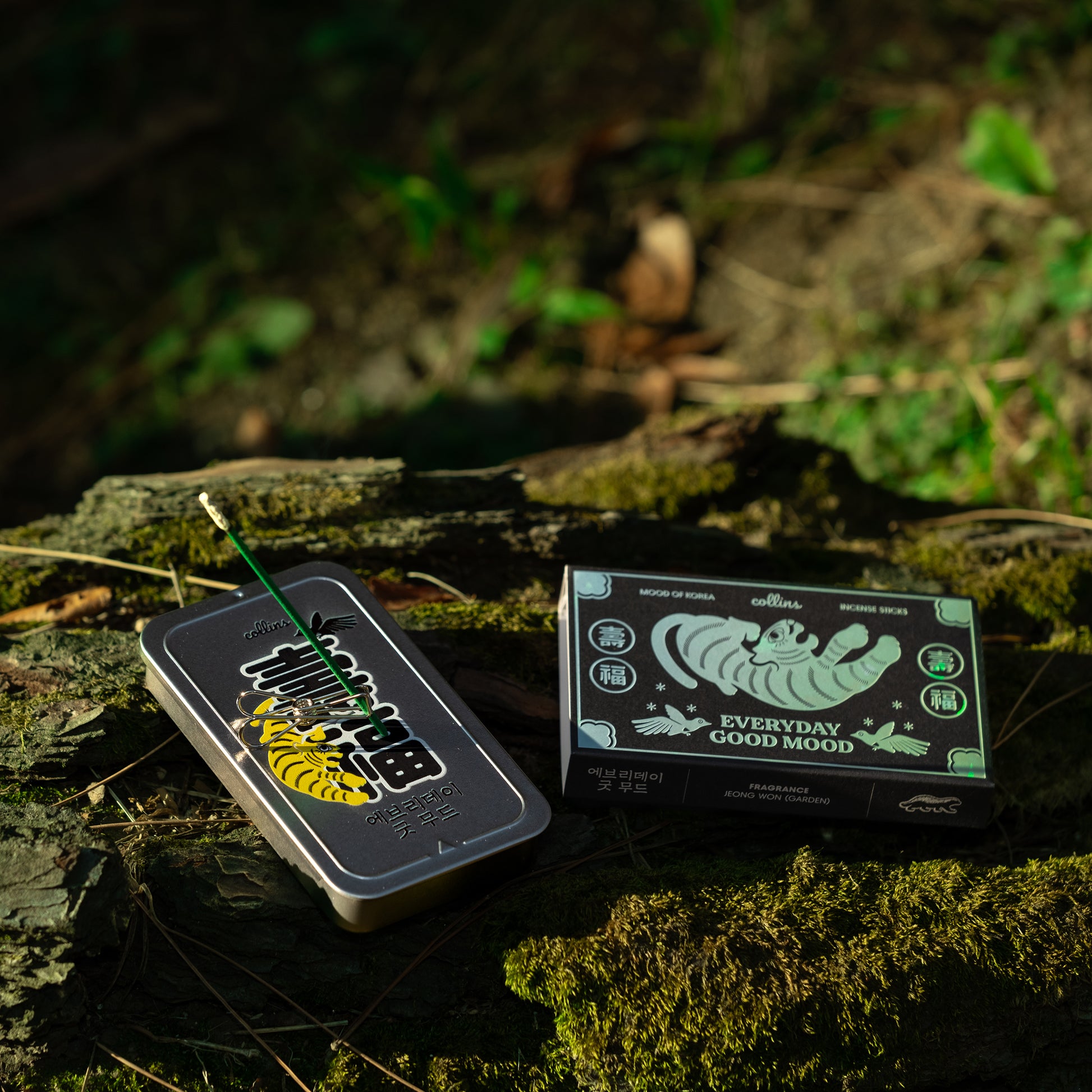 A tin of Collins natural incense sticks in korea edition scent, placed on a mossy log, with incense burning.