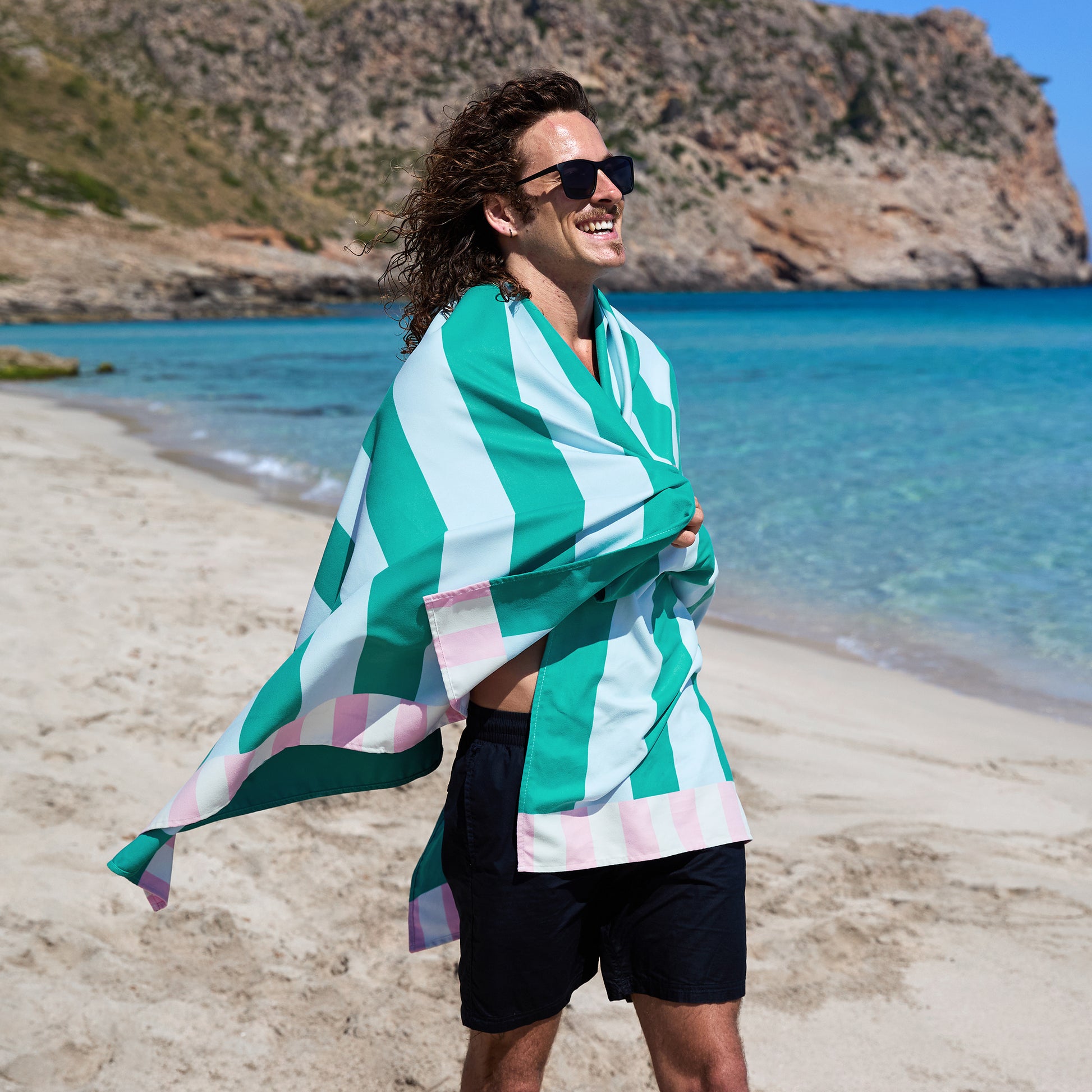 Showing a man on a beach wrapping him with Dock & Bay Beach Towel Clubhouse Collection XL Pastel Pier - oversized quick-dry beach towel with pastel multicolored stripes, 200x90cm, lightweight compact microfiber blend, sand-resistant, made from 100% recycled materials, includes handy pouch