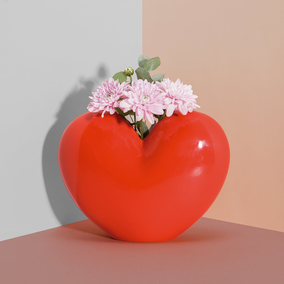 Red ceramic DOIY love vase filled with pale pink flowers sitting in corner of room on brown surface