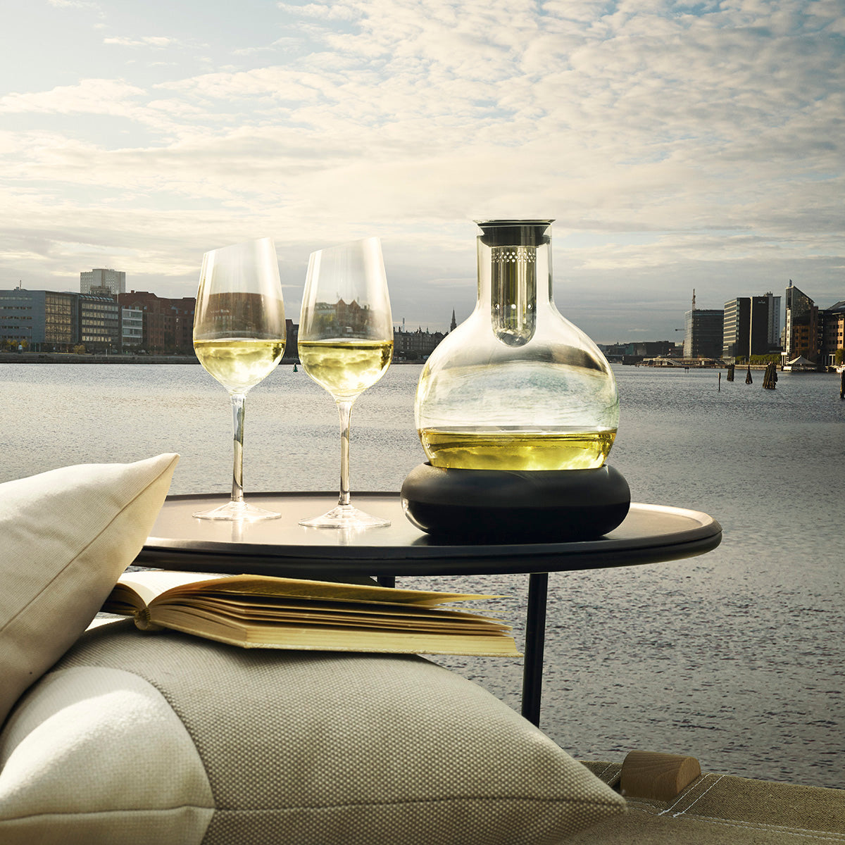 Bulb shaped Cool Wine Decanter by Eva Solo resting on it's cooling base filled with white wine with 2 glasses of wine and a view over the water in the background