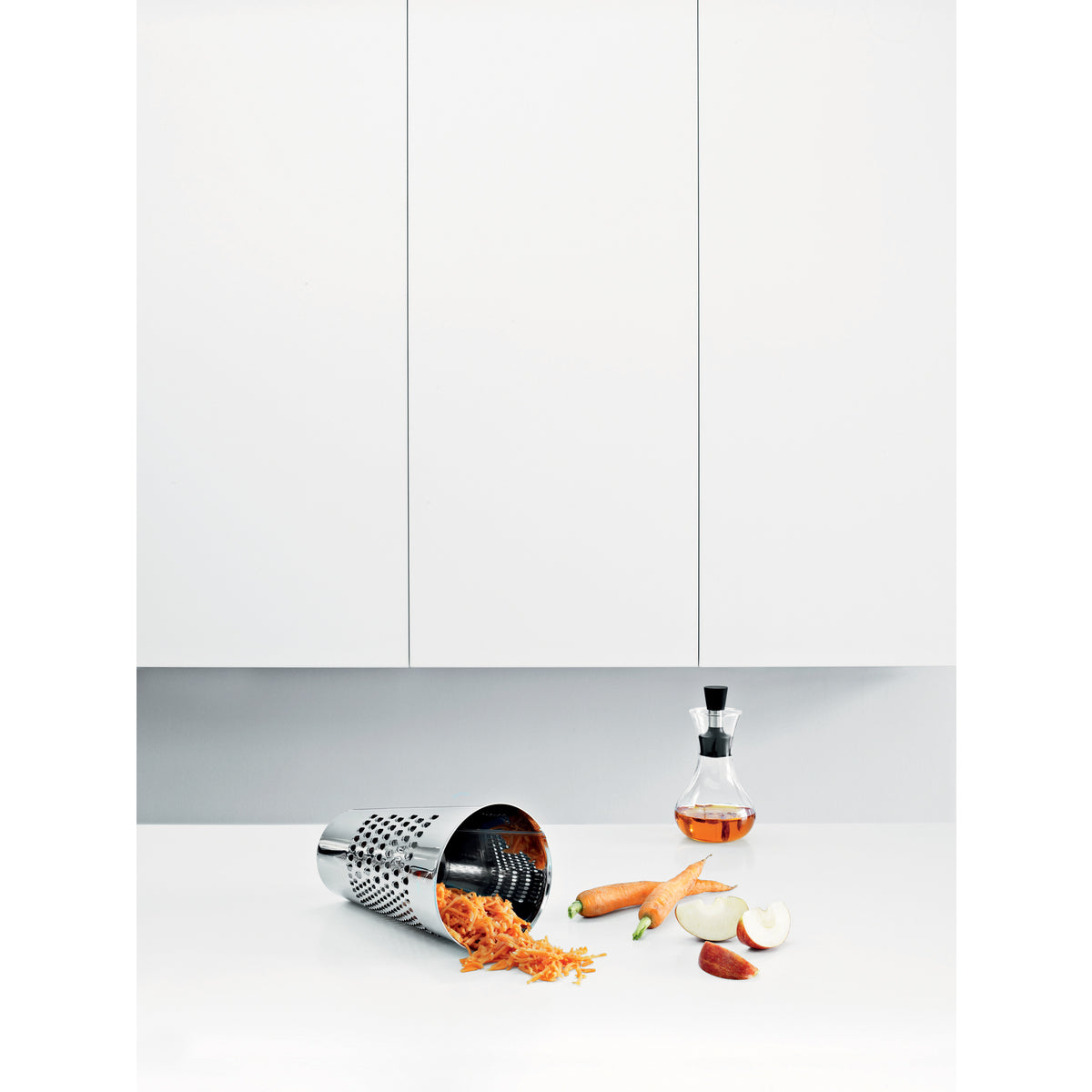 Laid on the table Eva Solo grating bucket with the grated carrots inside- stainless steel kitchen grater in silver color, cylindrical design for grating vegetables, cheese, and chocolate directly into the bowl.