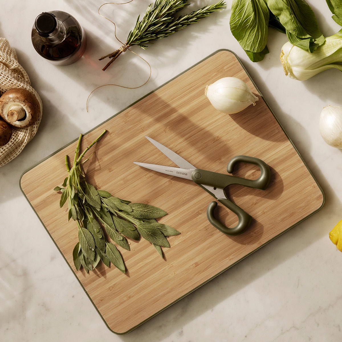 Green tool scissors by eva solo resting on a chopping board with herbs and vegetables.