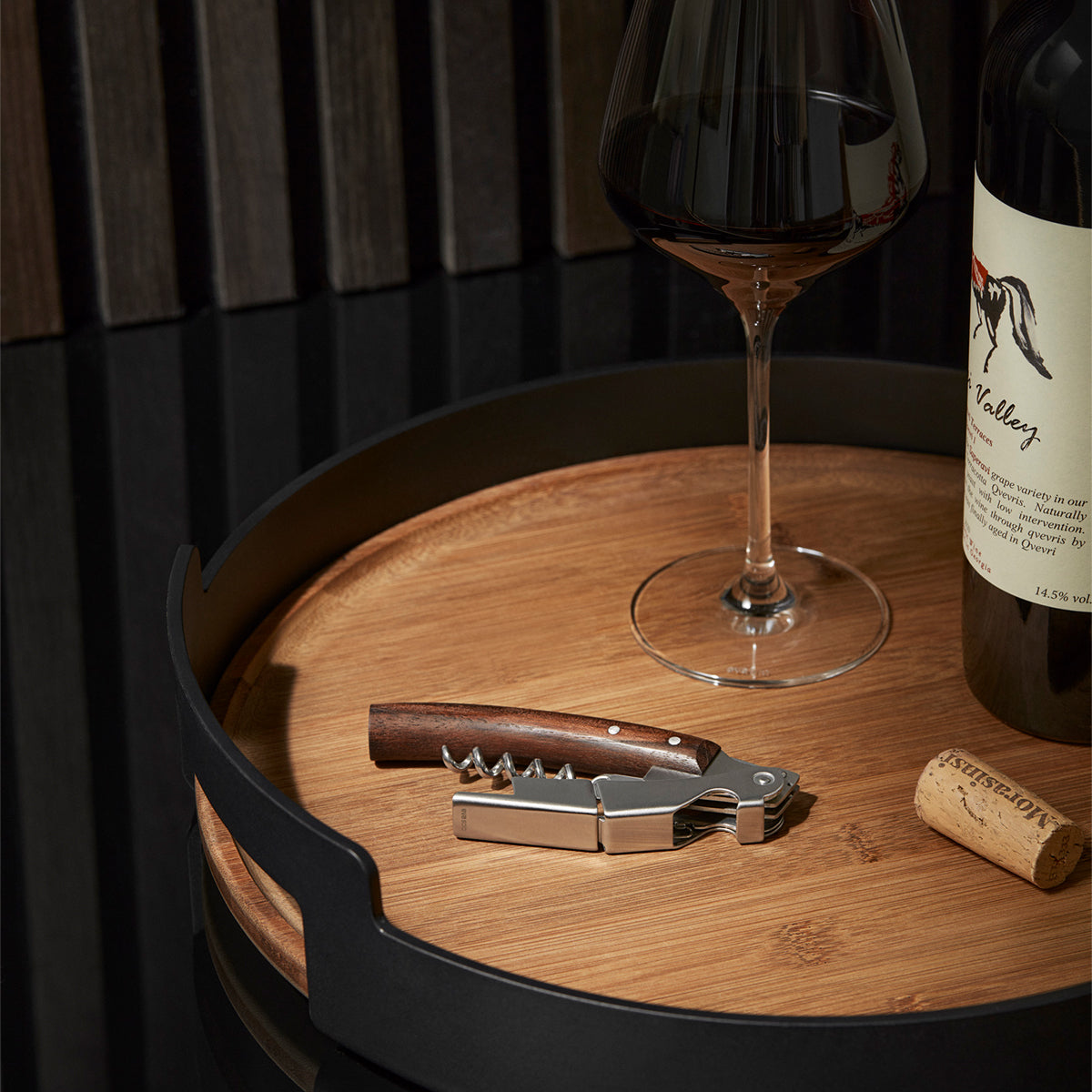Laid on the table with wine glass, wine and cork, Eva Solo Liquid Lounge waiter's corkscrew - brushed steel wine opener with oiled walnut handle, stainless steel worm, built-in foil cutter and bottle opener, compact two-step design.