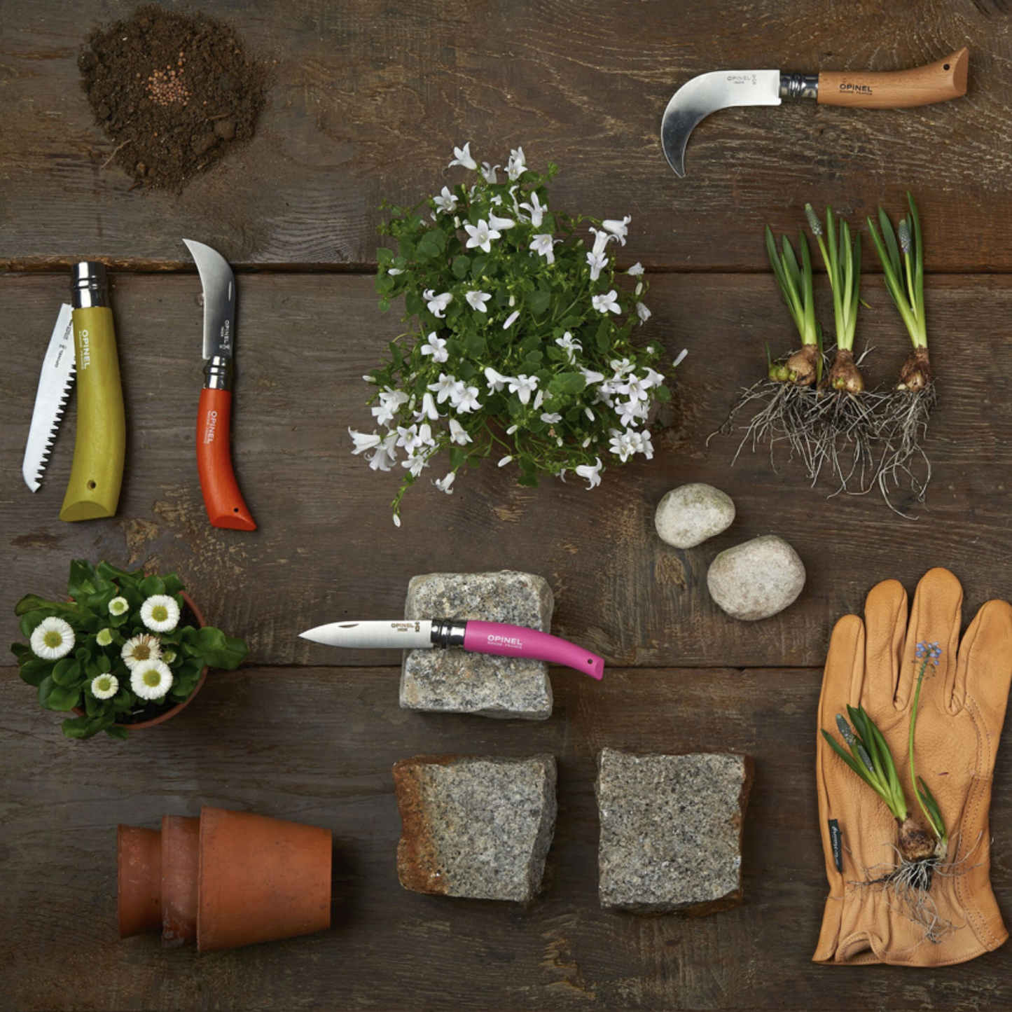 Opinel foldable Garden Tools 3pc Box Set on display with gardening gloves, soil, terracotta pots and flowers against wooden floor 