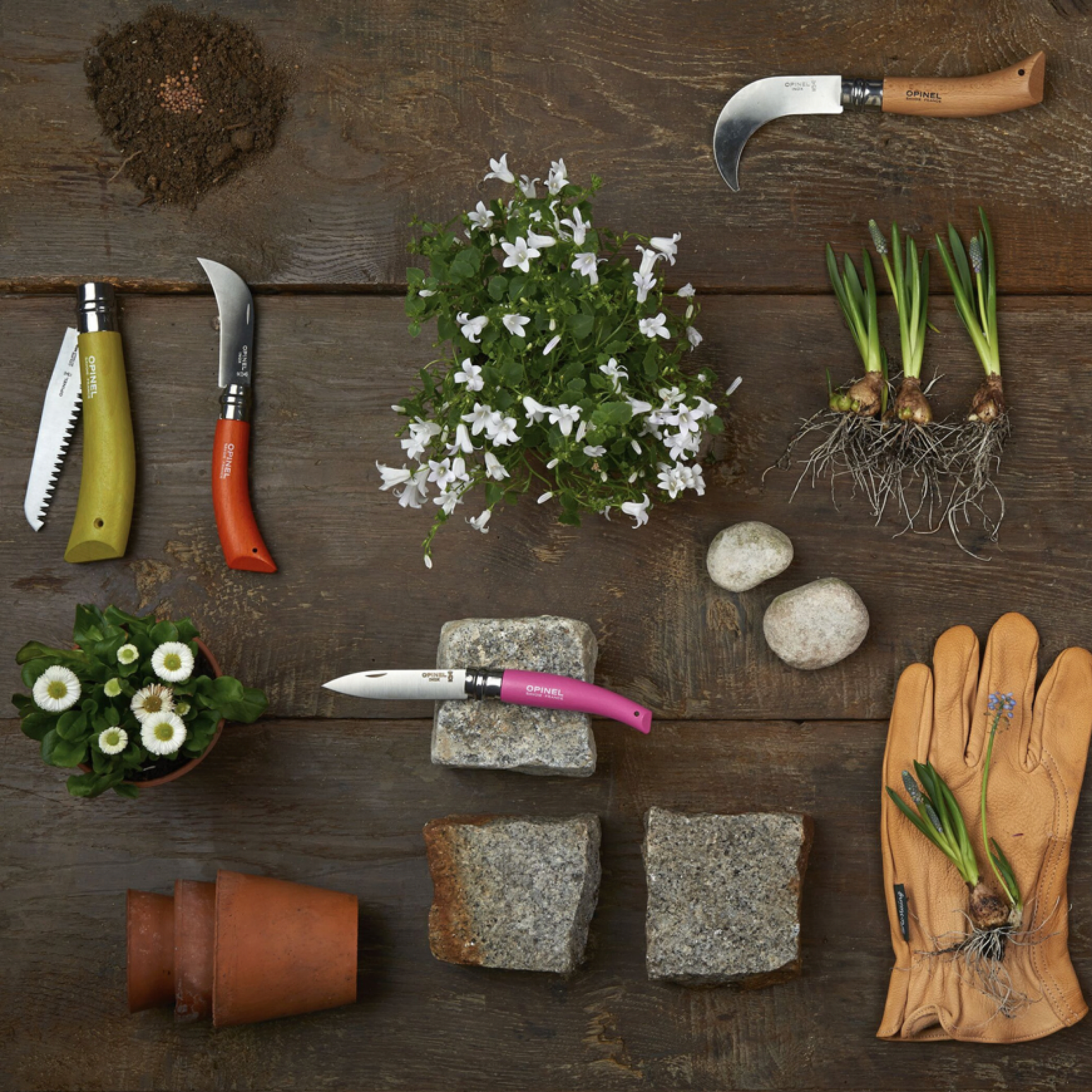 Opinel foldable Garden Tools 3pc Box Set on display with gardening gloves, soil, terracotta pots and flowers against wooden floor 