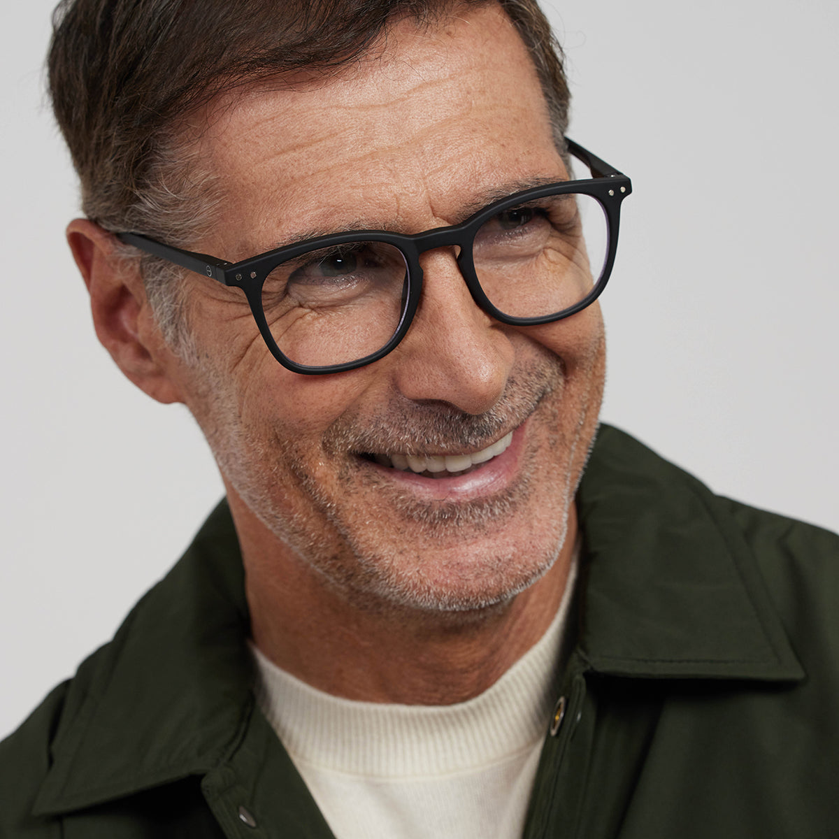 Man wearing Izipizi shape e black reading glasses, cream jumper under khaki shirt on an angle on white background
