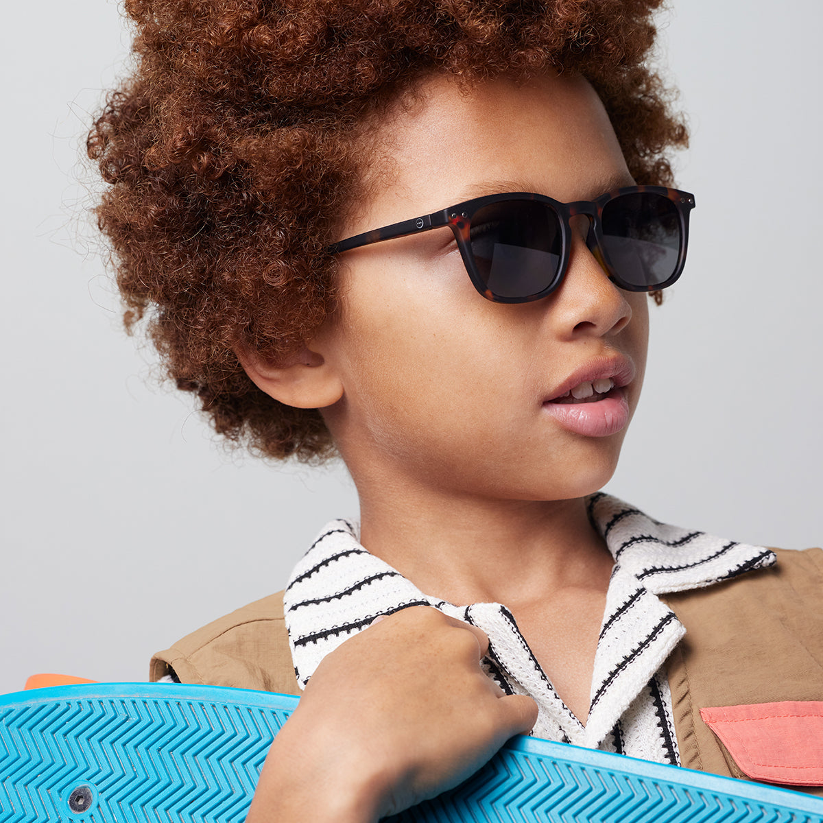 Boy with brown curly hair and white striped top wearing Izipizi junior sunglasses shape e tortoise holding blue skateboard