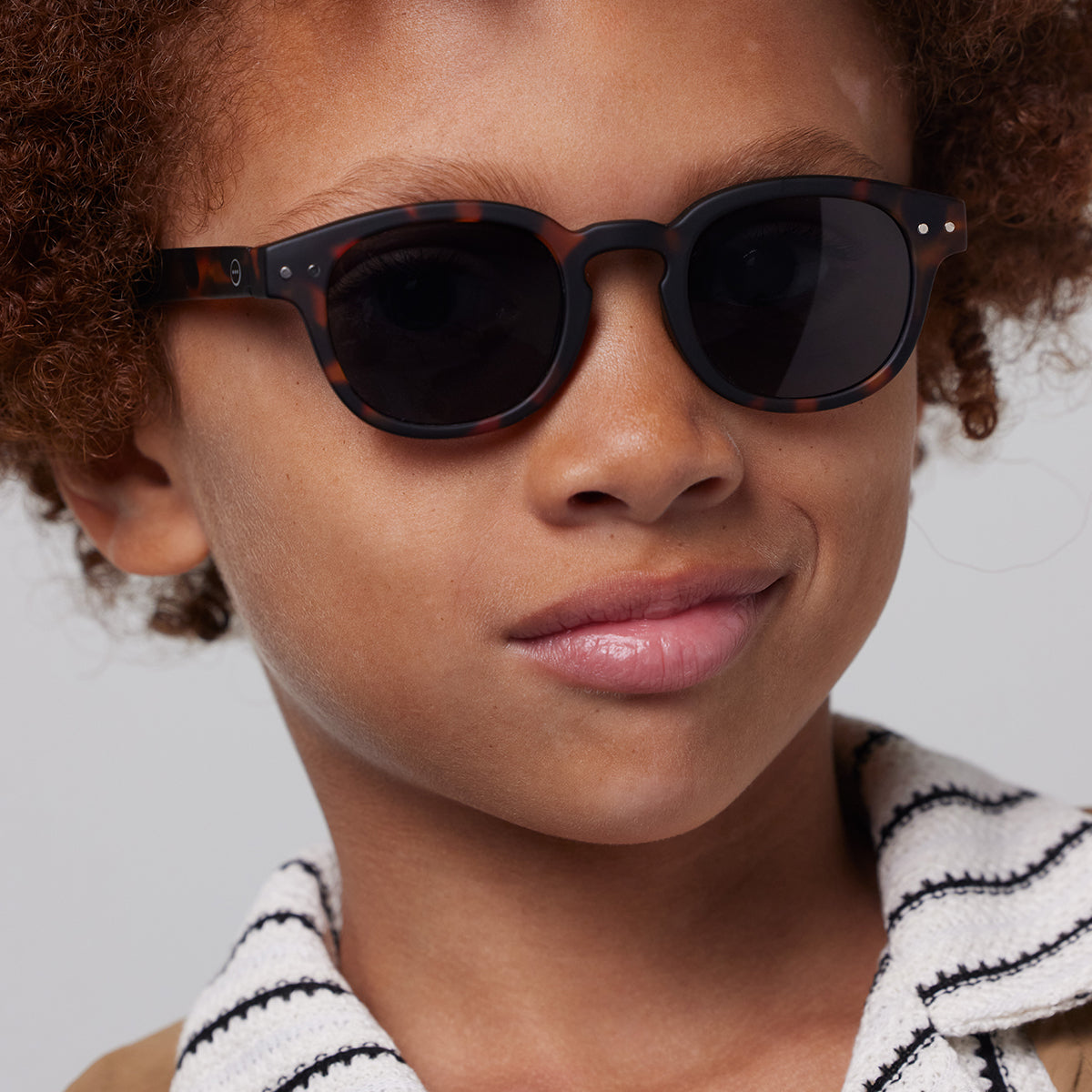 Boy with brown curly hair and white striped top wearing Izipizi junior sunglasses shape c tortoise 