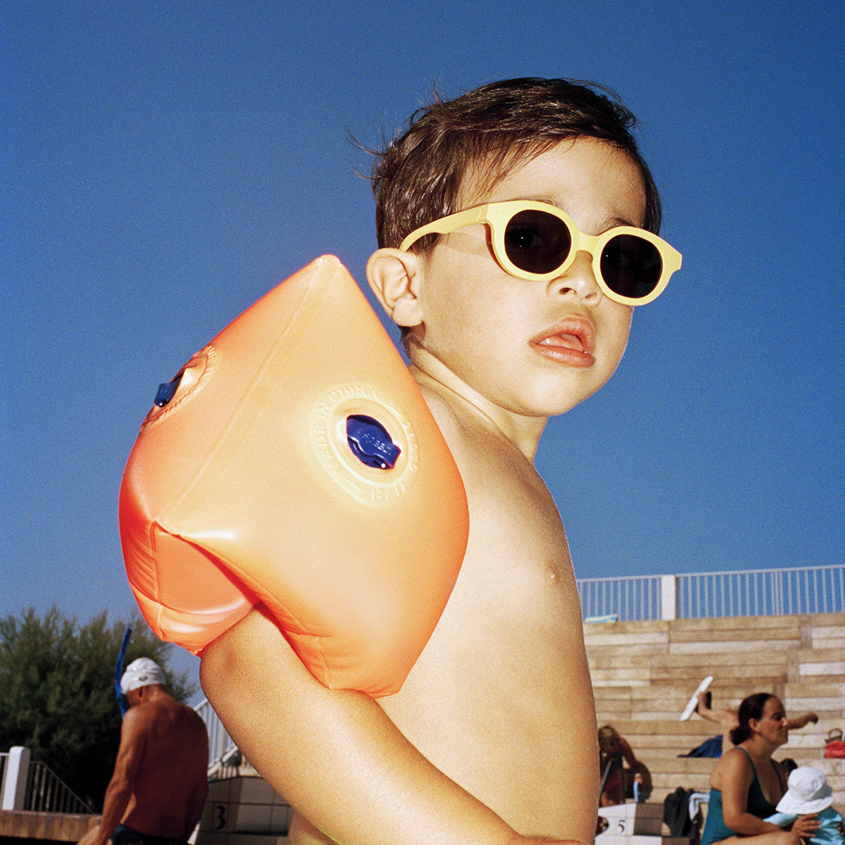 Little boy wearing orange arm floats at the beach wearing Izipizi kids sunglasses shape c lemonade 