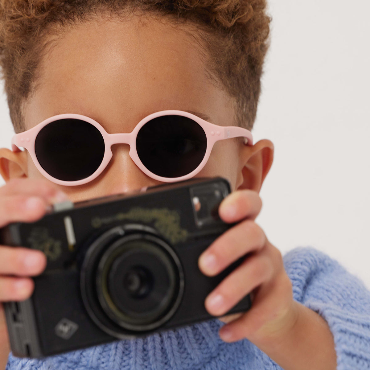 Little boy in blue knitted jumper with camera wearing Izipizi kids sunglasses shape d pastel pink