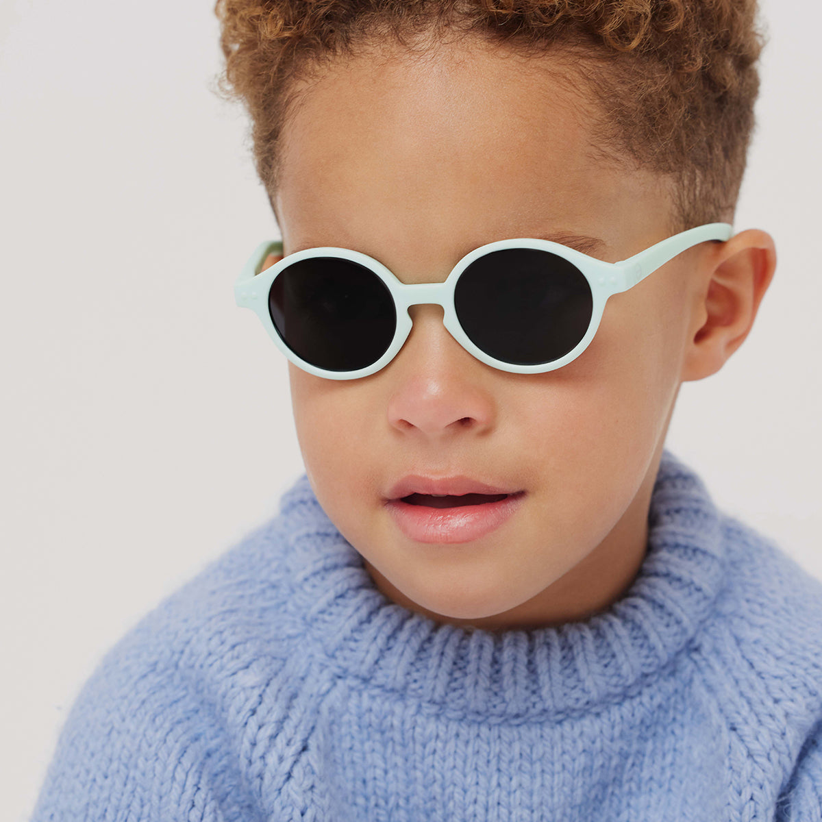 Little boy in blue knitted jumper wearing Izipizi kids sunglasses shape d sweet blue