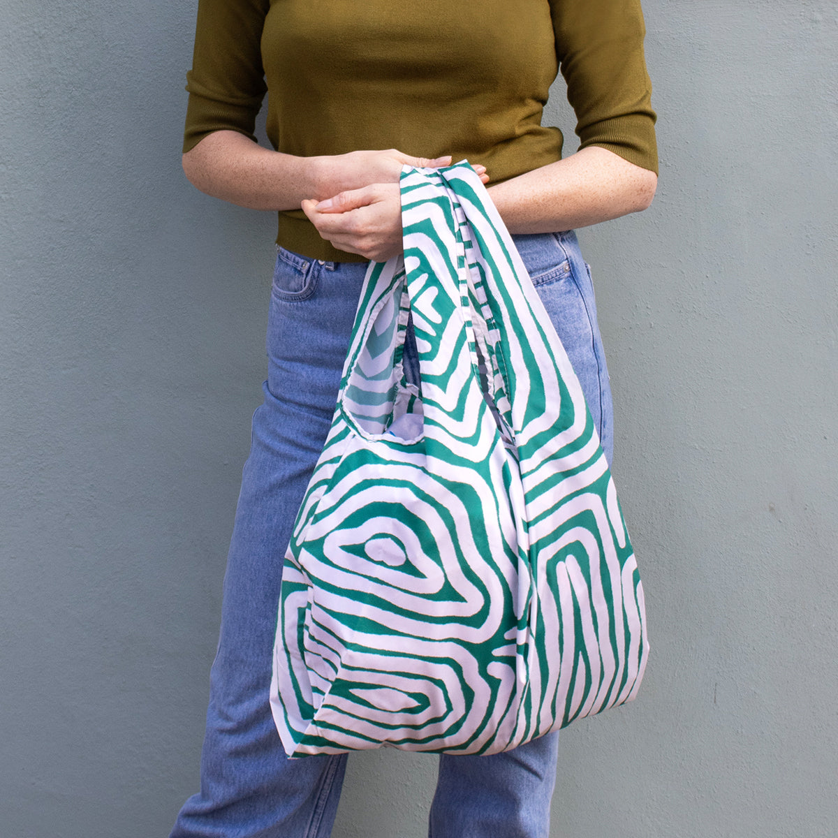Image showing Regusable bag by Kind bag in green and white abstracted lines print, shown slung over a woman's arm.