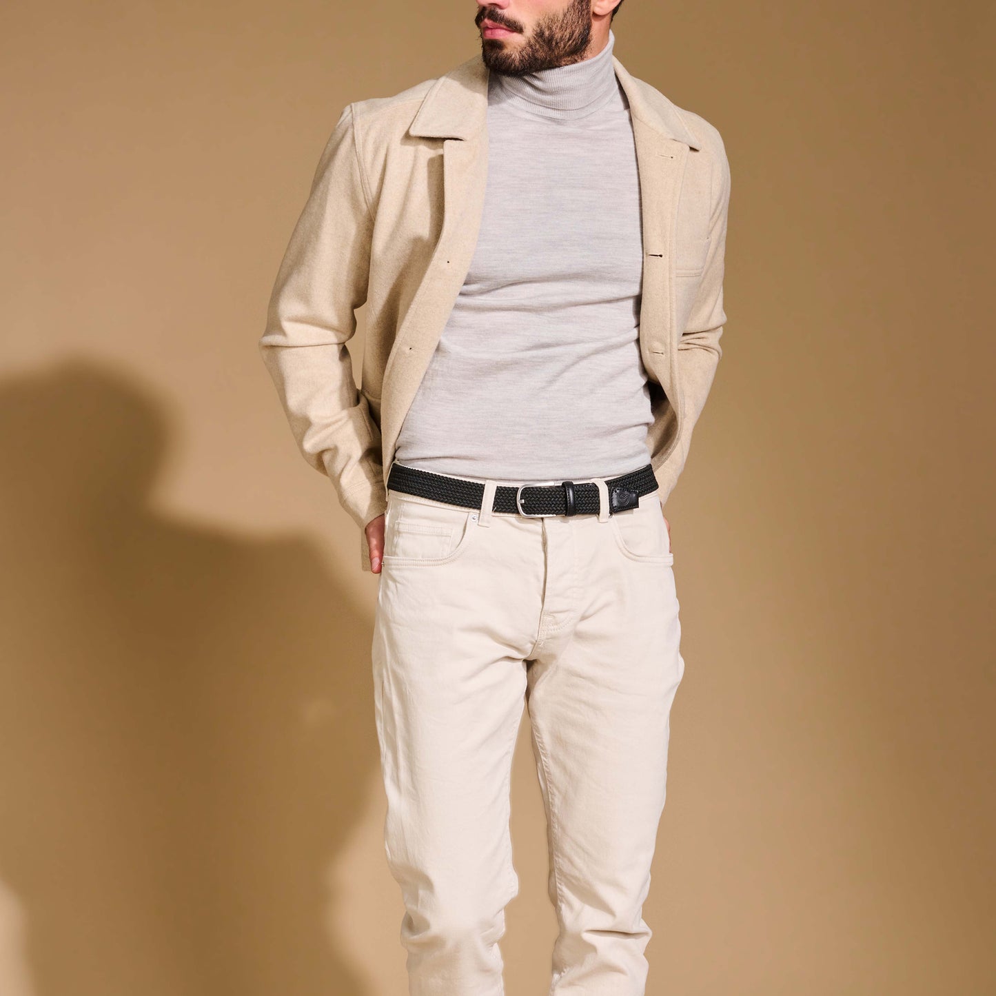Close up of man wearing La Boucle mono London woven belt around off white pants, beige jacket and grey polo top on brown background