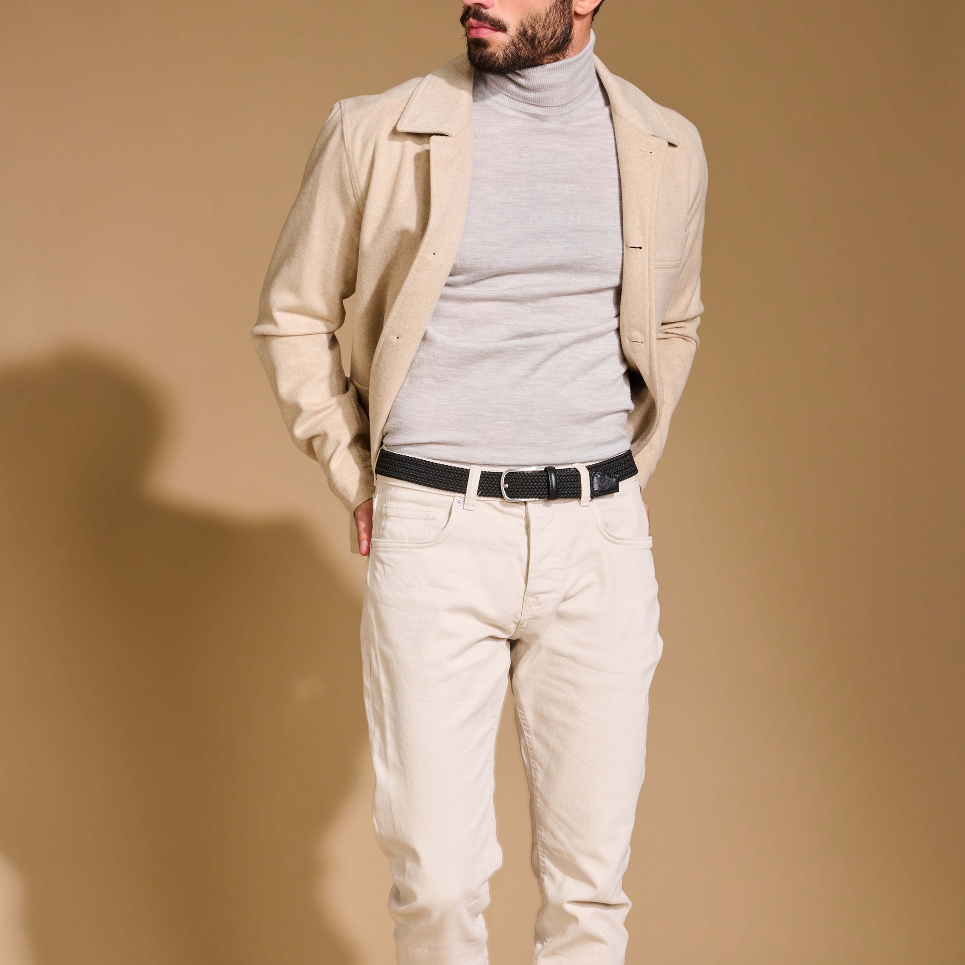 Close up of man wearing La Boucle mono London woven belt around off white pants, beige jacket and grey polo top on brown background