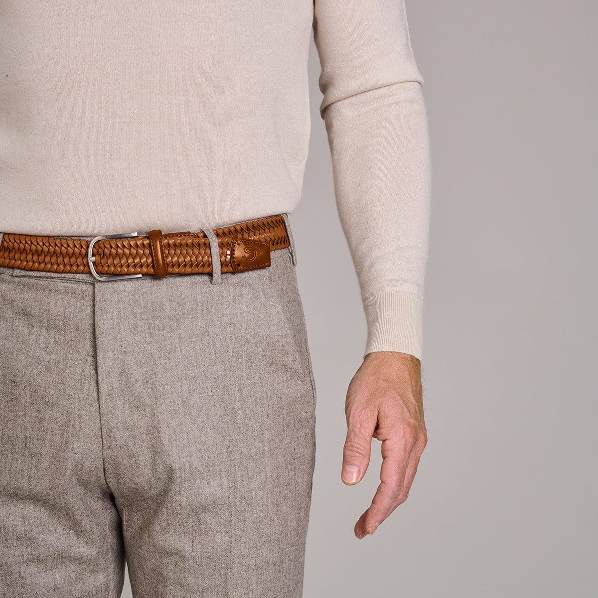 Close up of man wearing La Boucle stretch leather mono Napoli woven belt around grey pants and long sleeve cream top on grey background