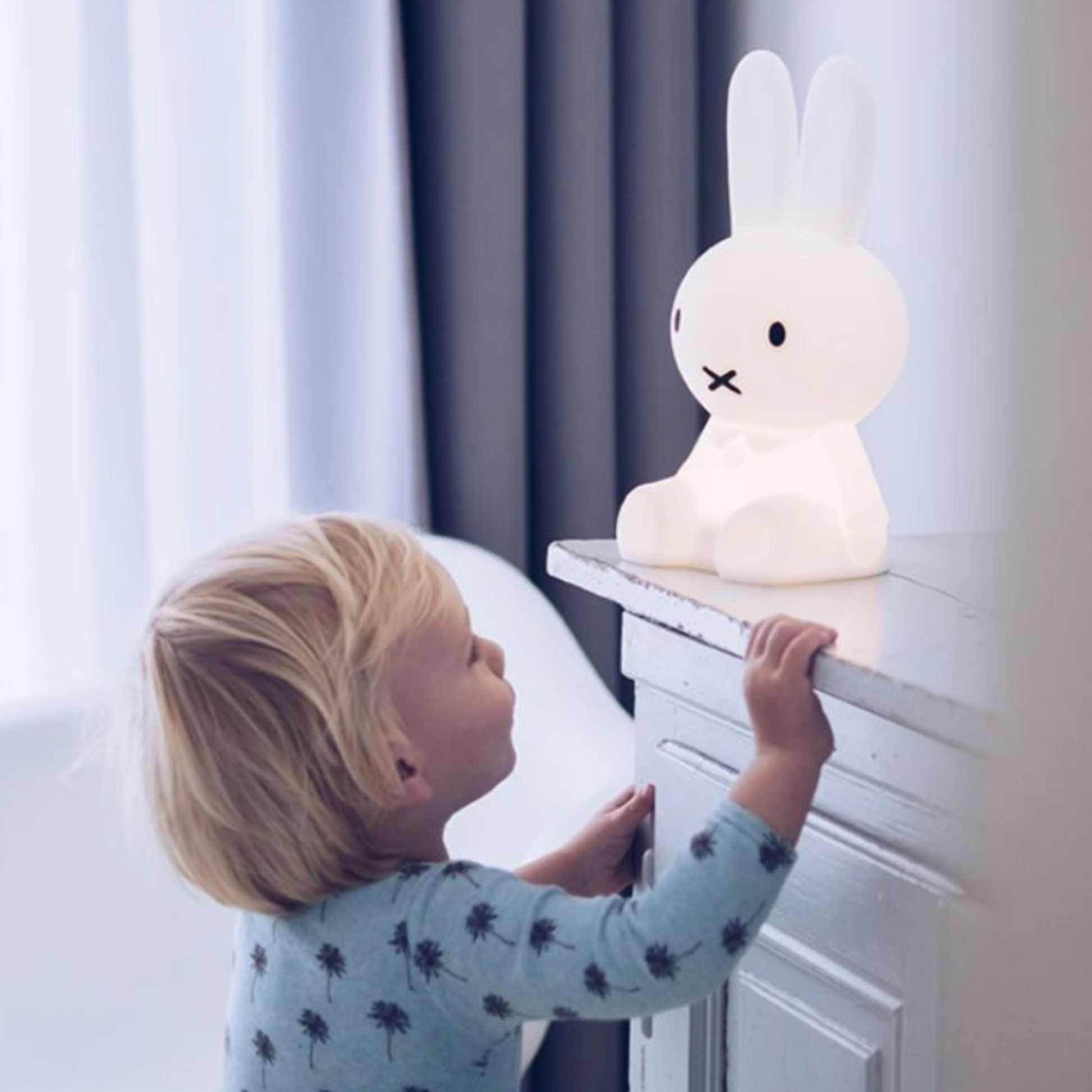 Lifestyle shot of childrens Mr Maria white Miffy 30cm night light on display in childs room on mantlepiece turned on with child peering up at light