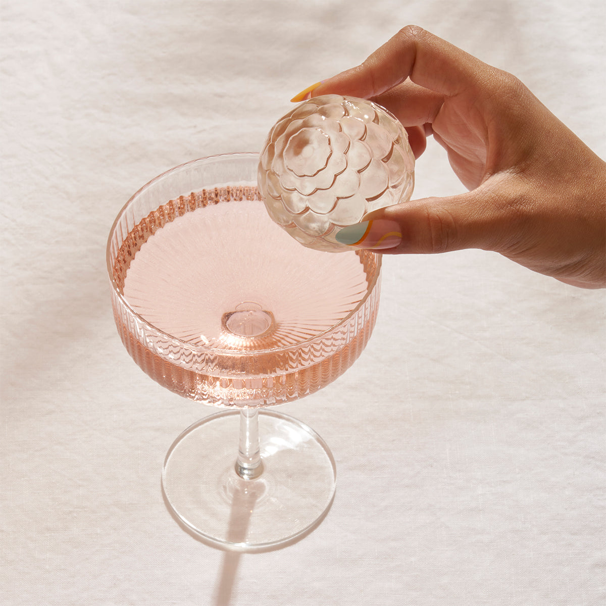 WP charcoal petal cocktail ice cube held above rippled cocktail glass with a pale pink drink