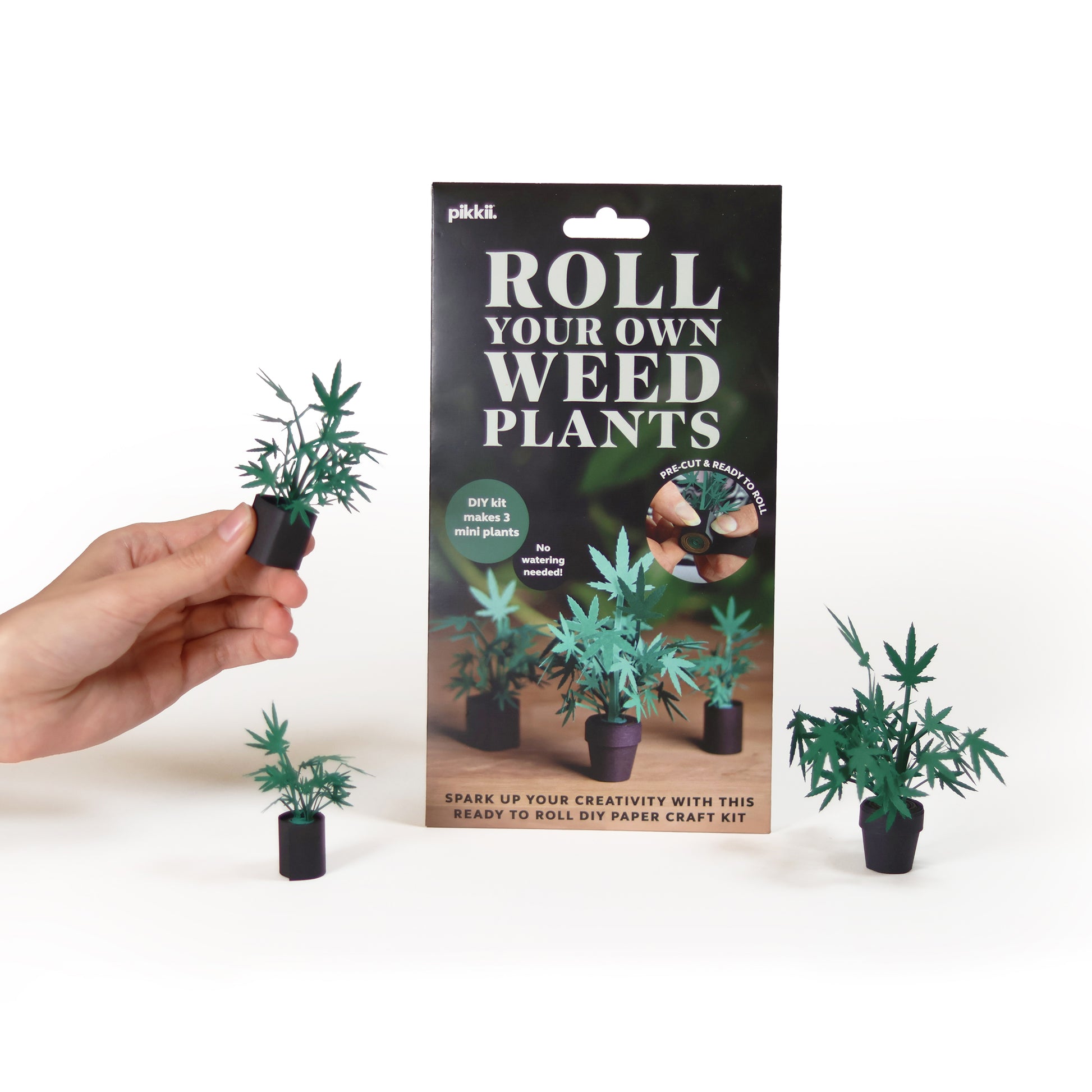 Three paper marijuana plants and the packaging from Pikkii Roll Your Own Weed Plants paper craft kit - premium FSC-paper DIY kit with pre-cut sheets for creating three paper marijuana plants, plastic-free packaging, soy-ink printing, 140×260×5 mm size, includes step-by-step instructions.