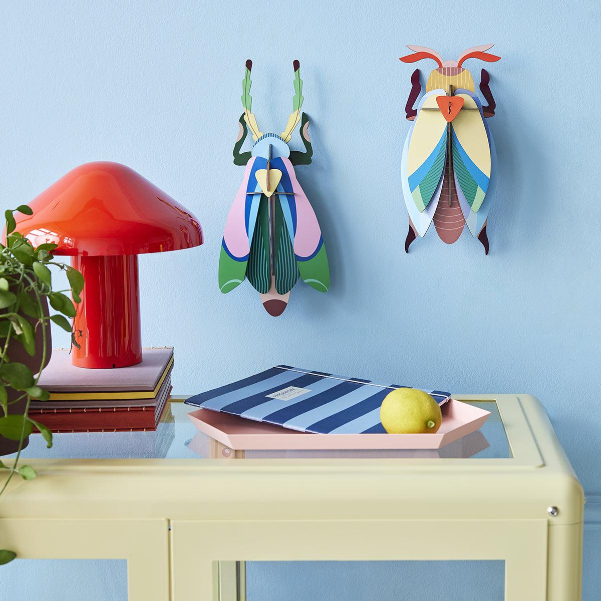 Studio Roof A4 lunar jewel beetle wall art and one other buy on a pale blue wall in front of a lemon desk with red lamp and lemon