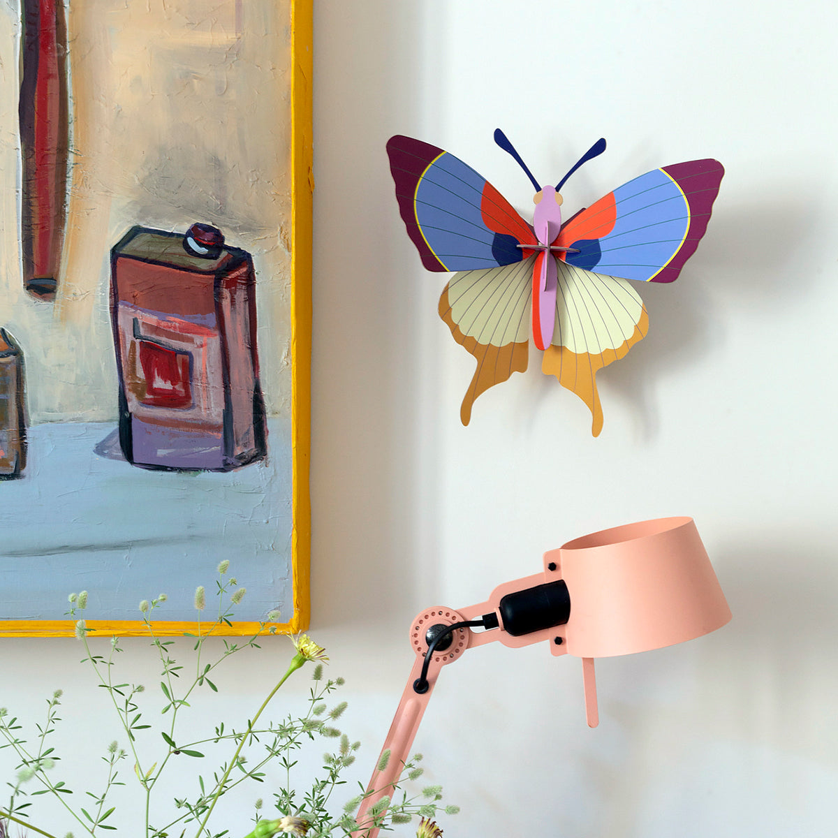 Studio Roof A4 plum fringe butterfly wall art mounted on white wall next to painting with yellow frame and peach lamp below 