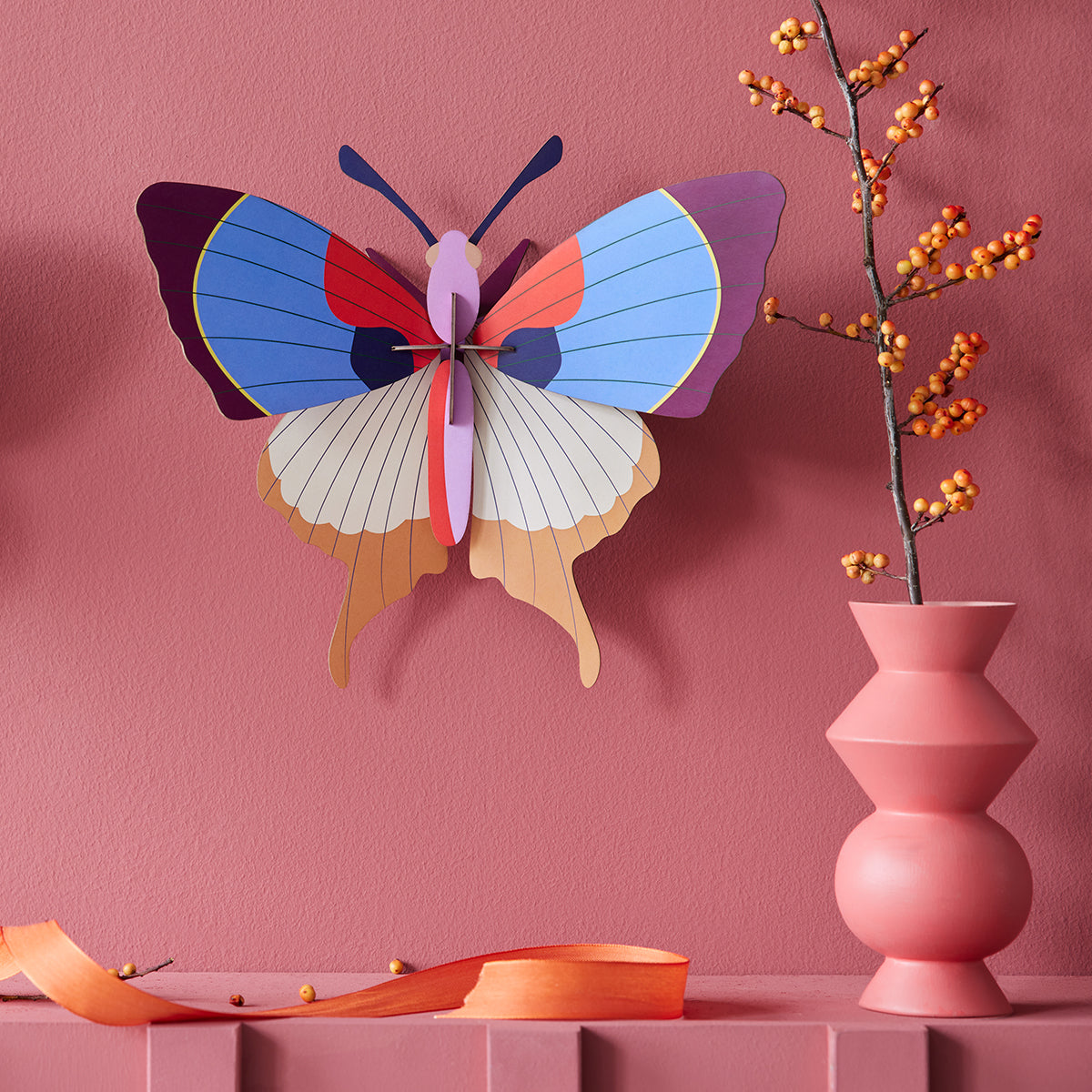 Studio Roof A4 plum fringe butterfly wall art on dark pink wall next to pink vase with orange seed display